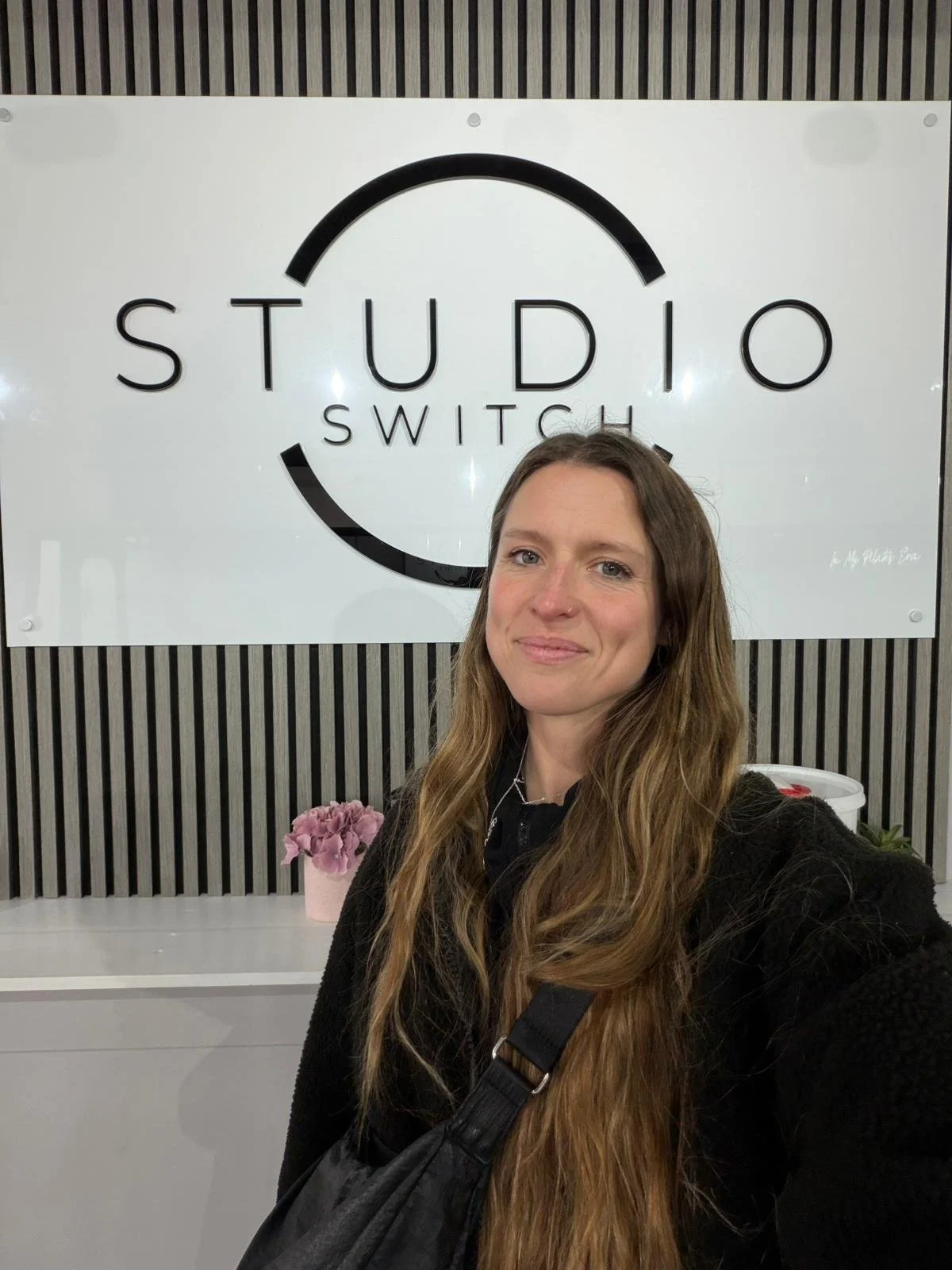 A woman with long wavy hair taking a selfie in front of a sign that says "Studio Switch" with a flower in a pink pot on a white counter.