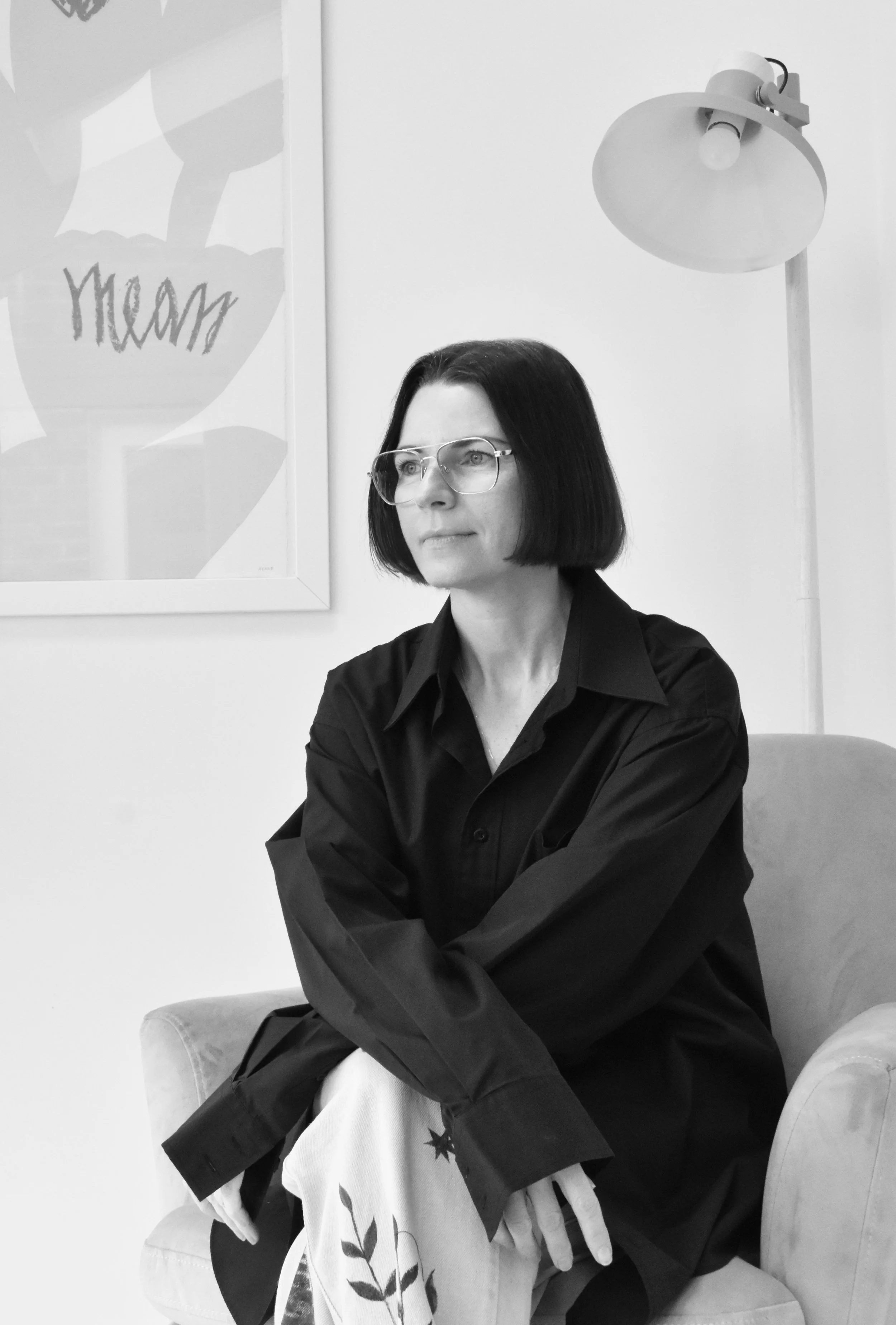 Black and white photo of a woman with glasses seated on a chair, wearing a black shirt and holding a patterned cloth. A lamp and a framed abstract artwork are in the background.