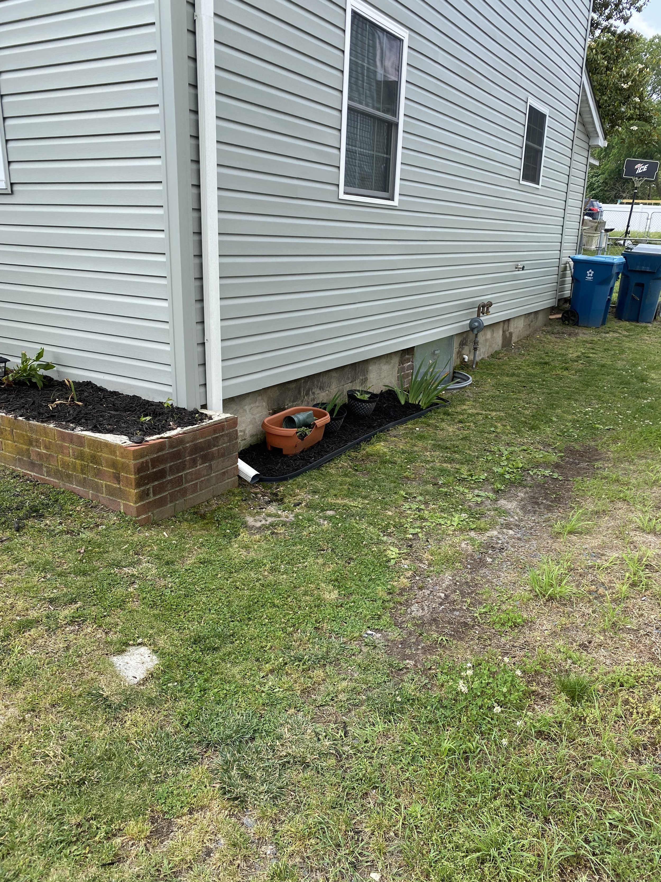 Side view of a house with light gray vinyl siding, two windows, and a brick planter. There are potted plants, a garden hose, black mulch, and green grass. Blue recycling and trash bins are visible in the background.