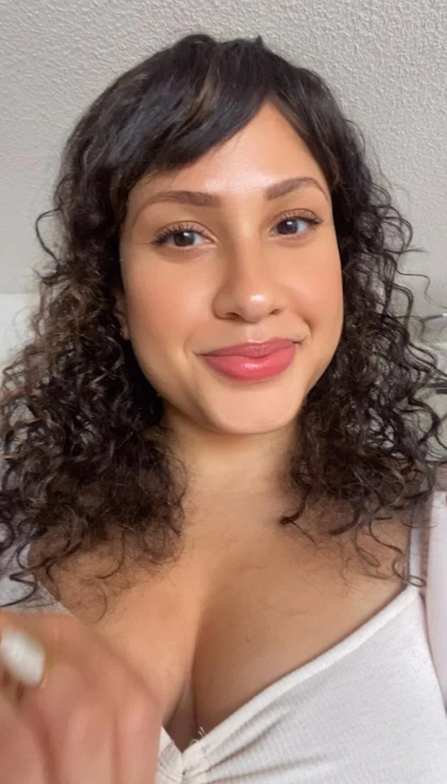Close-up of a woman with curly dark hair, smiling, wearing a white top, against a plain background.