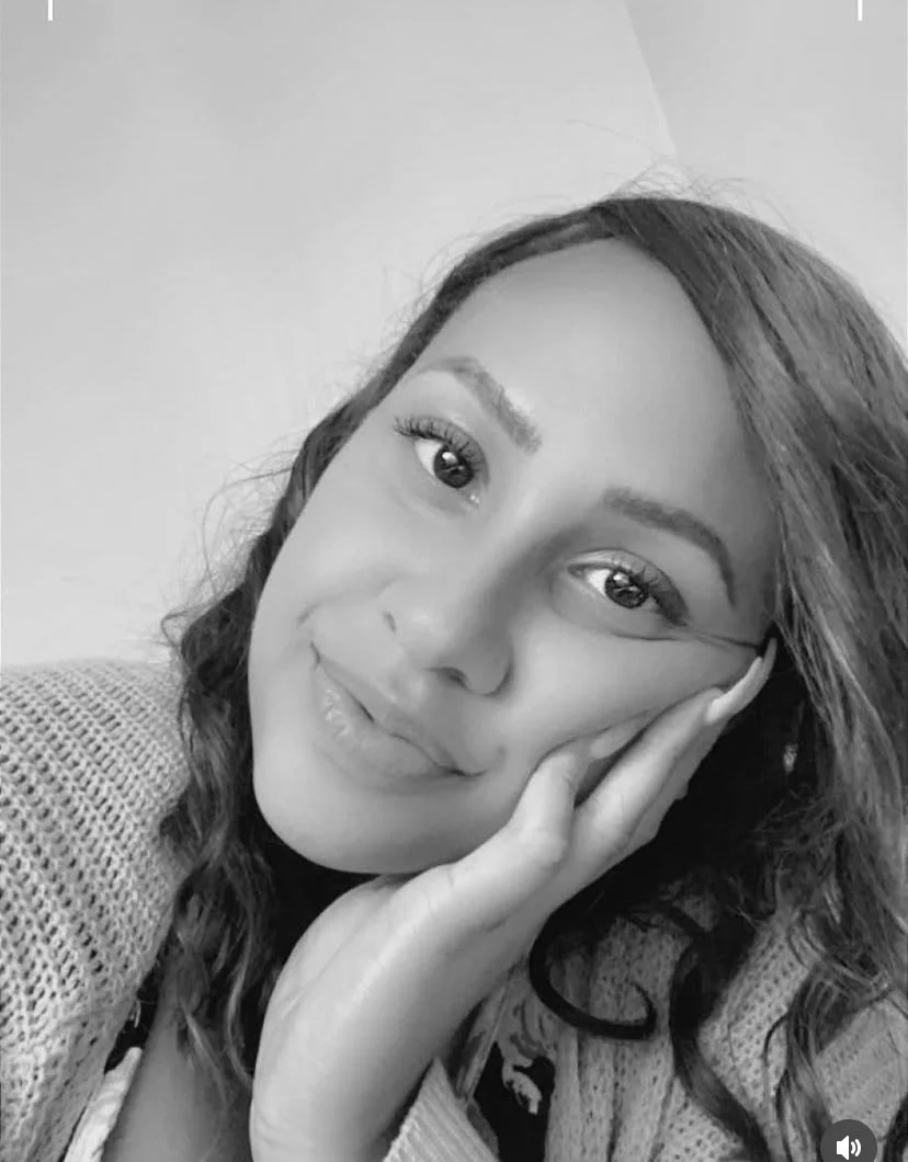 A black and white close-up photo of a woman resting her face on her hand, smiling softly, with wavy hair and wearing a textured sweater.