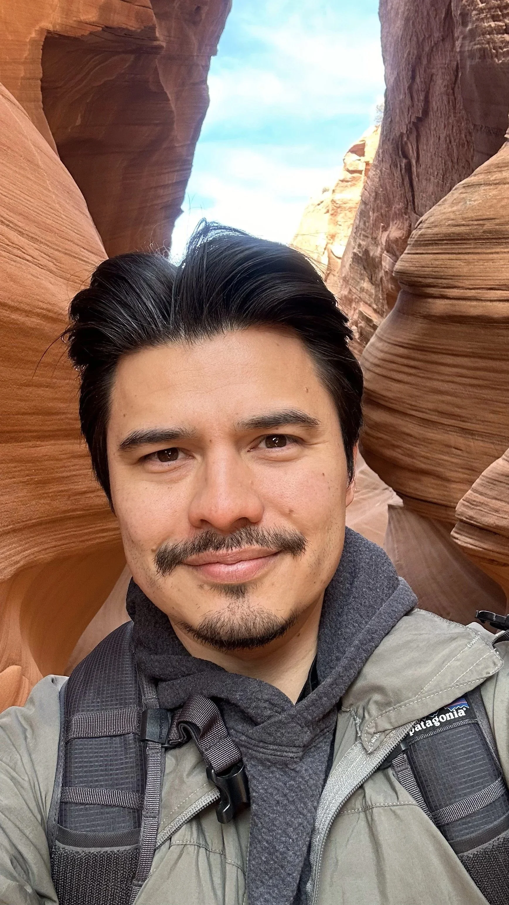 A man taking a selfie in a canyon with smooth, layered sandstone walls and a blue sky with some clouds in the background.