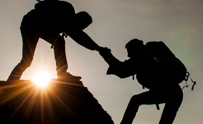 Two hikers silhouetted against the sky, helping each other climb a mountain during sunset.