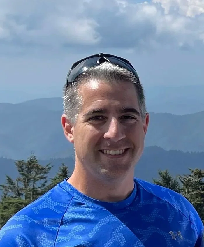 A man smiling outdoors with mountains and trees in the background, wearing a blue athletic shirt and sunglasses on his head.