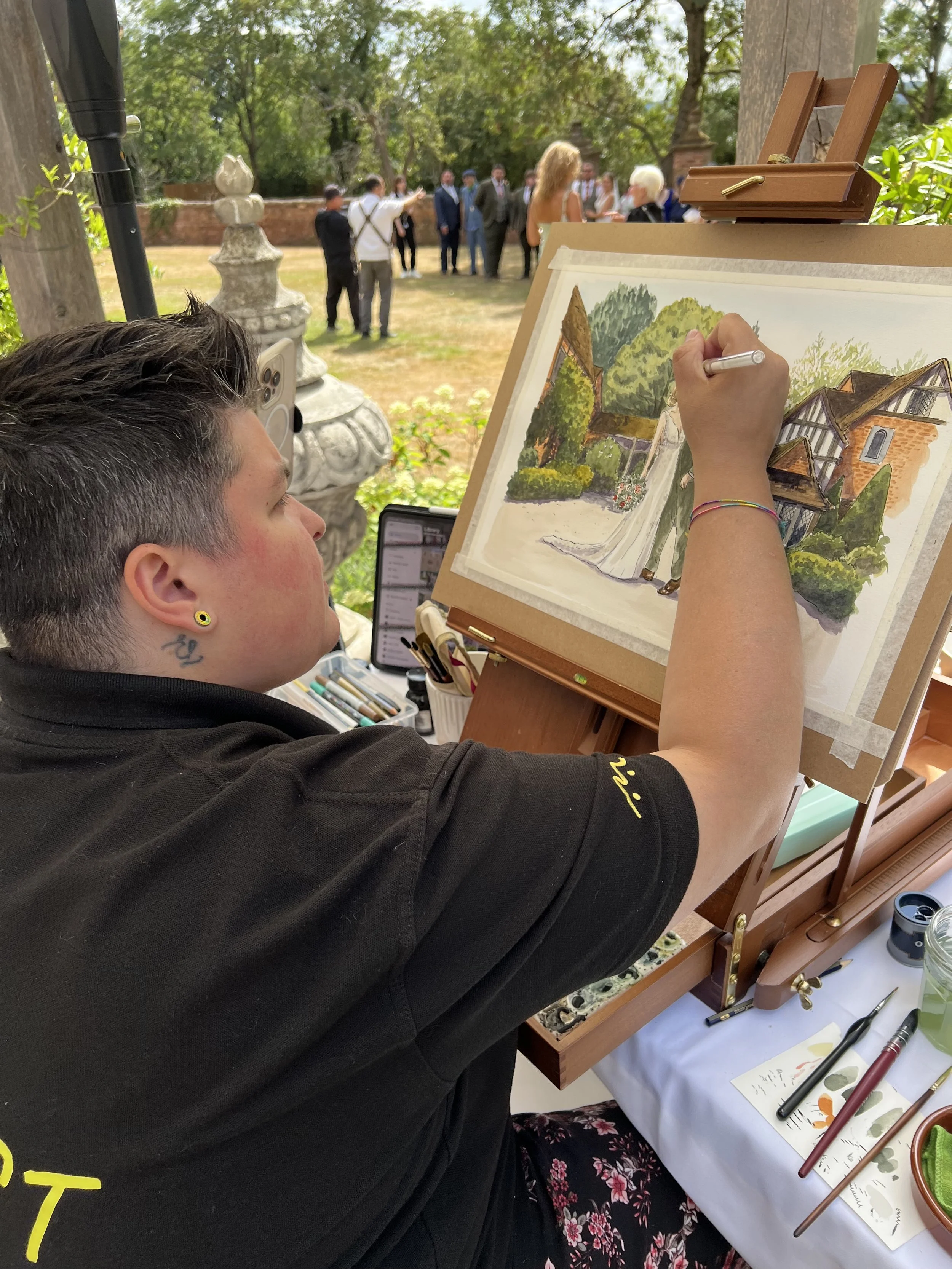 An artist creating a watercolor painting of a garden scene with a woman and a building, sitting at an outdoor art station with brushes and supplies.