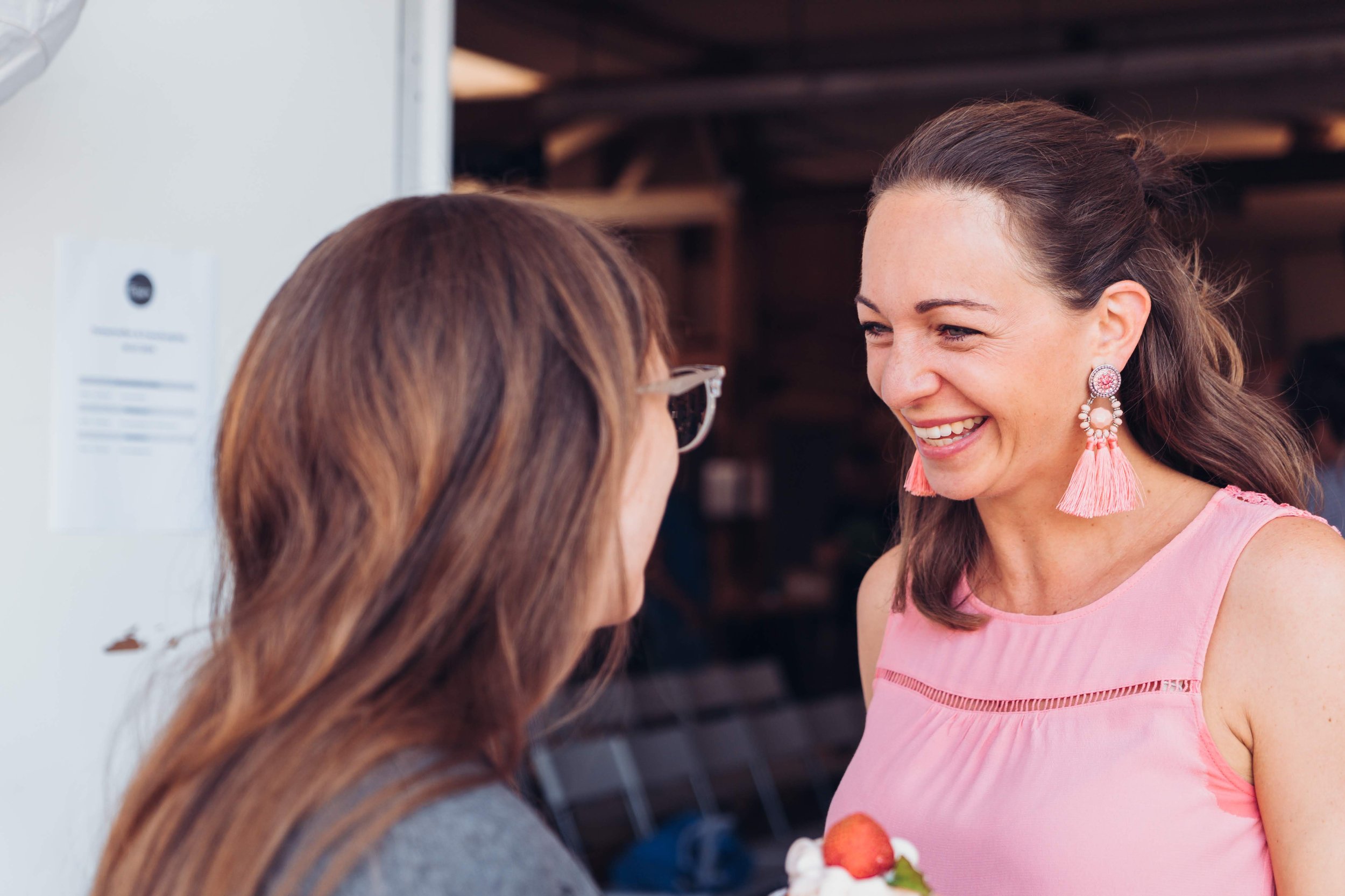 Zwei Frauen lachen sich fröhlich an, eine hält einen Teller mit einem Erdbeerkuchen, bei einem Treffen im Freien.