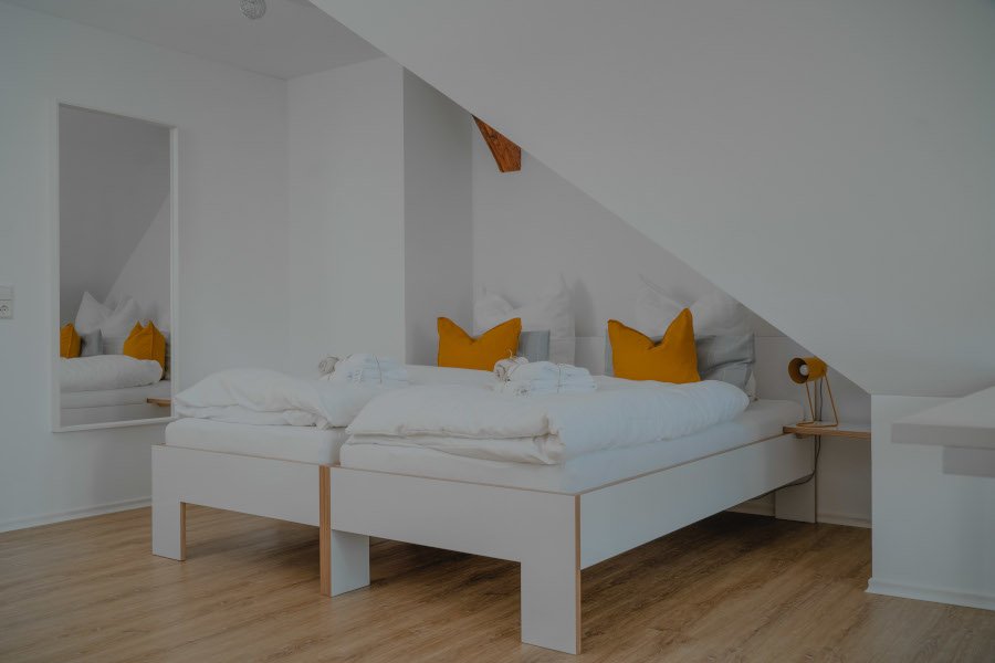 Ein Schlafzimmer mit weißen Möbeln, einem Bett mit weißen und gelben Kissen, einem Wandspiegel und einem an der Wand befestigten Nachttisch mit einer Lampe.