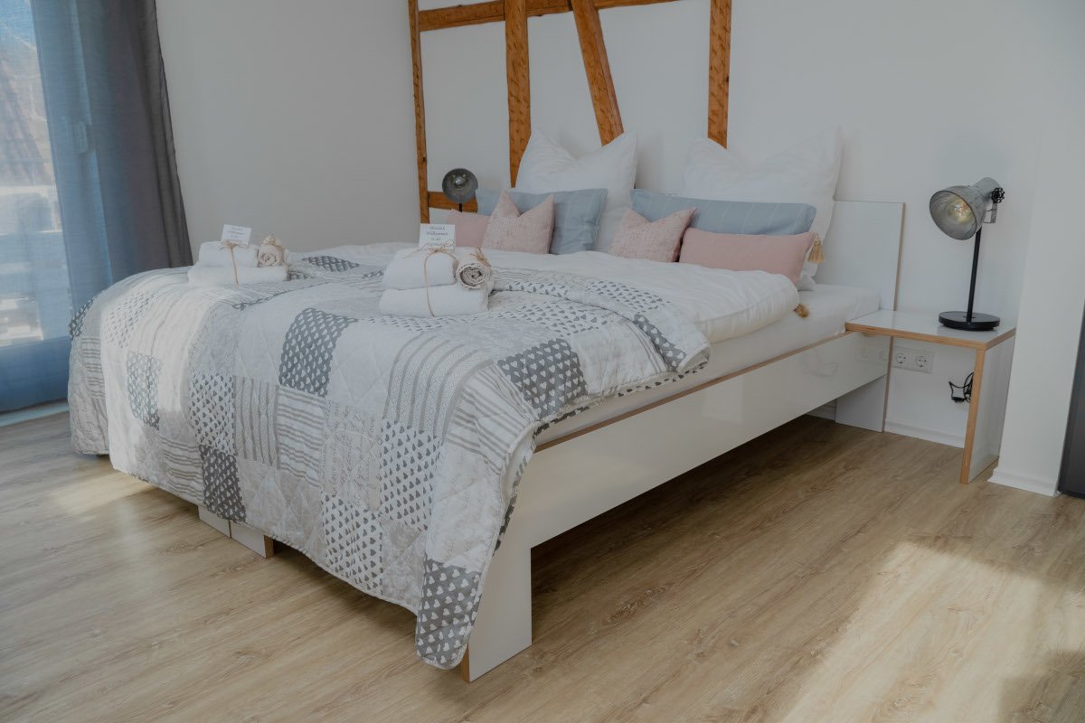Ein bequemes Bett mit weißen, grauen und pastellfarbenen Kissen sowie Handtüchern auf Bettwäsche mitgrauen und weißen Mustern, in einem modernen Schlafzimmer mit Holzlamellenkopfteil, seitlichem Nachttisch mit Lampe, auf hellem Holzboden.