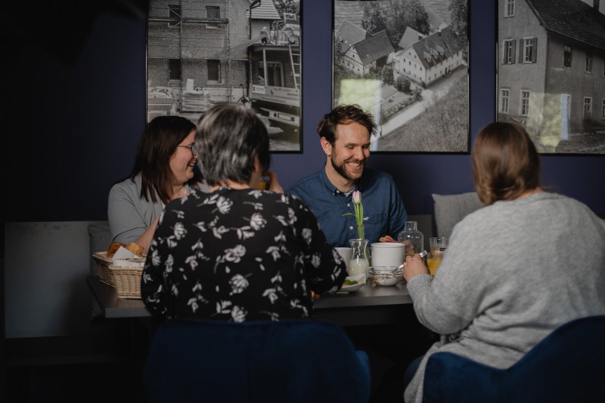 Vier Personen sitzen an einem Esstisch in einem Restaurant und unterhalten sich. Auf dem Tisch sind Teller, Gläser und eine Vase mit einer Tulpe. Im Hintergrund sind vier Wandbilder mit Schwarz-Weiß-Fotografien von Gebäuden.