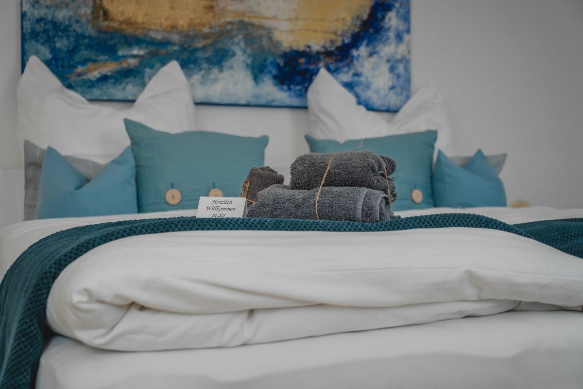 Ein ordentlich gedecktes Hotelbett mit weißen und blauen Kissen, einem blauen Bettläufer, handtuchrollen und einem Willkommensschild, im Hintergrund ein abstraktes Gemälde.
