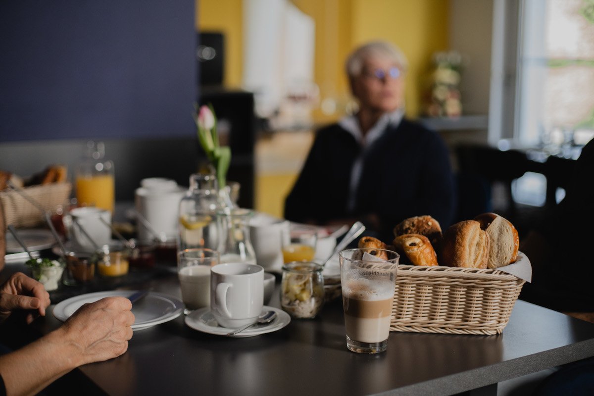 Frühstückstisch mit Croissants, Brötchen, Kaffee, Orangensaft und Joghurt, im Hintergrund eine ältere Frau mit Brille am Fenster.