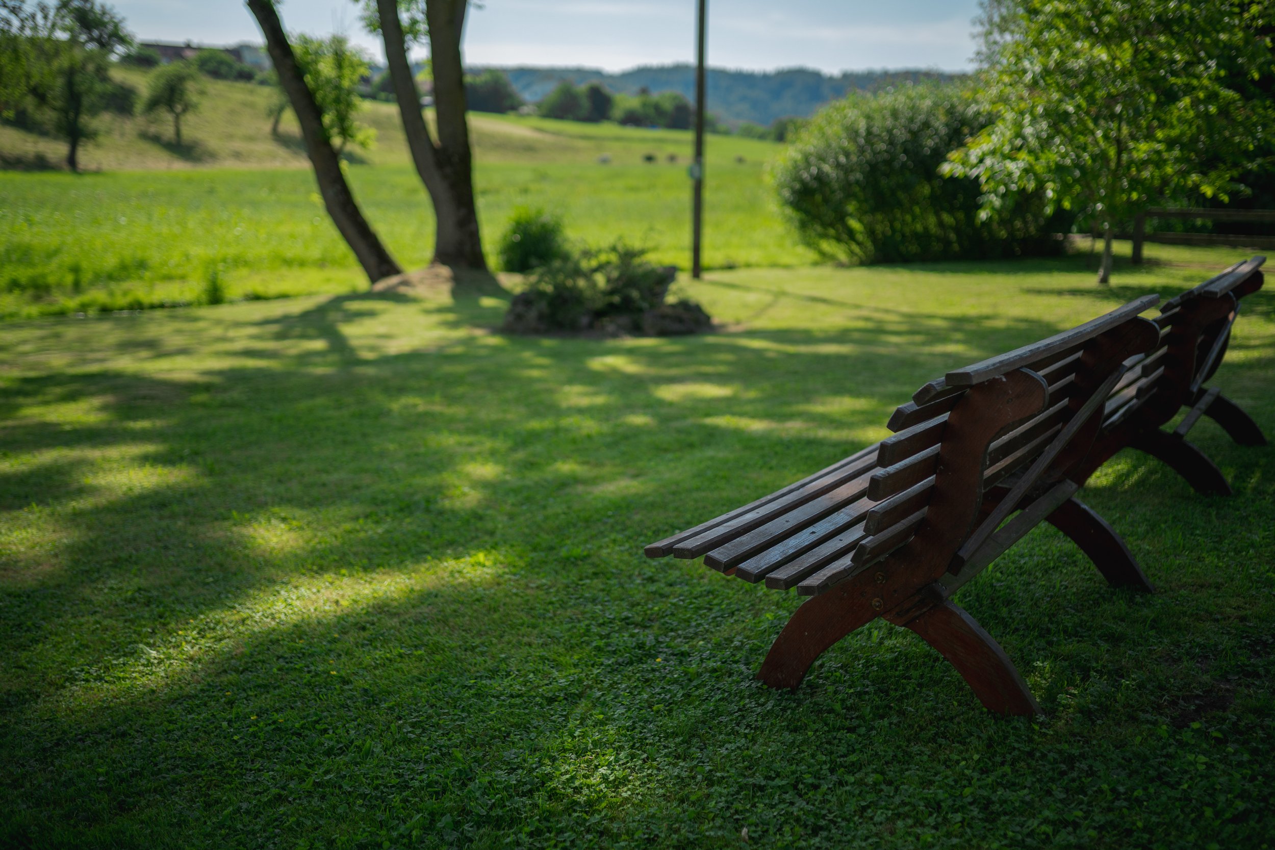 Zwei Holzbänke auf einer Rasenfläche in einem parkähnlichen Garten, im Hintergrund Bäume und eine weite grüne Landschaft unter einem blauen Himmel.