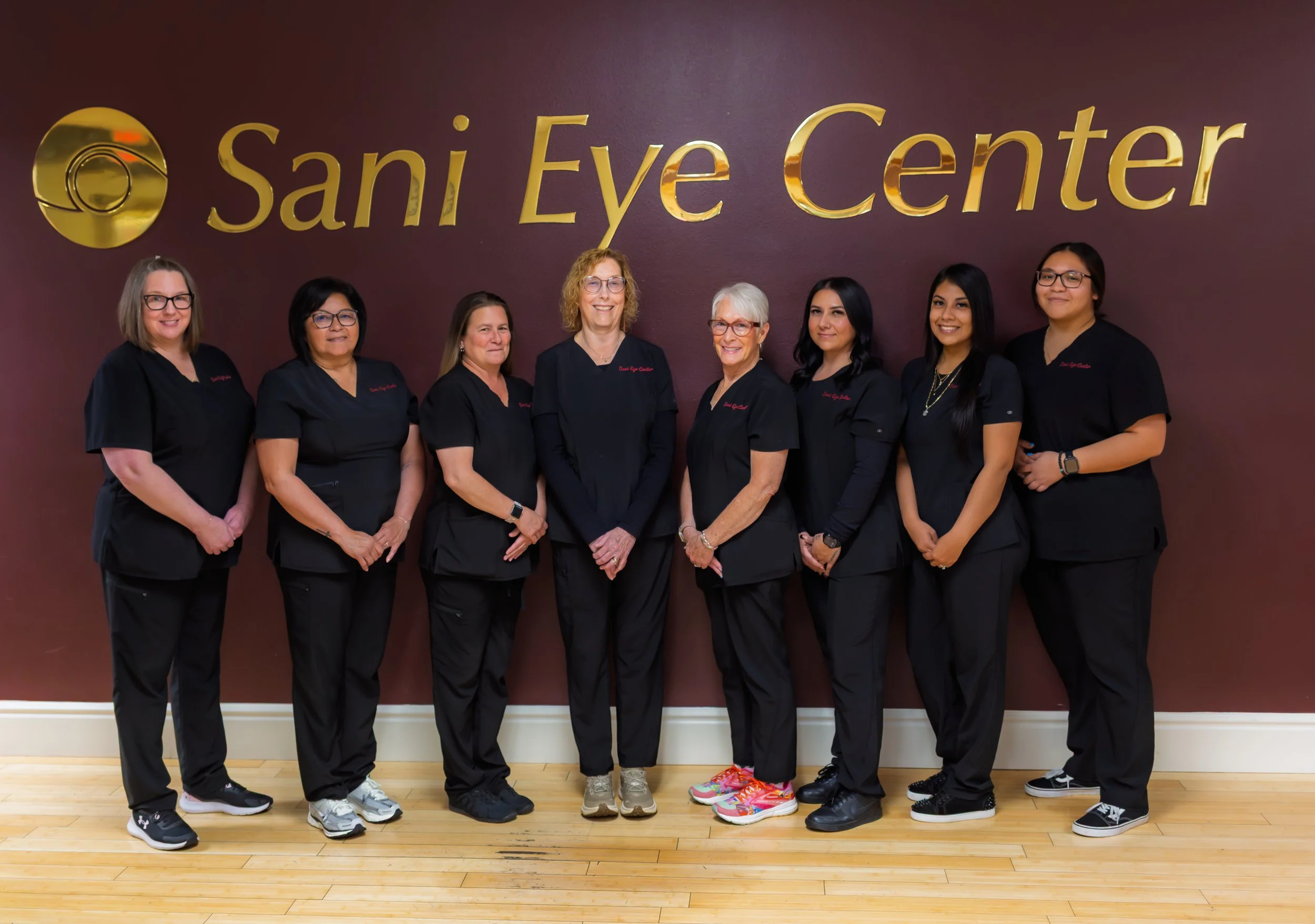 Group of women dressed in black scrubs standing in front of a maroon wall with the gold sign 'Sani Eye Center' and logo.