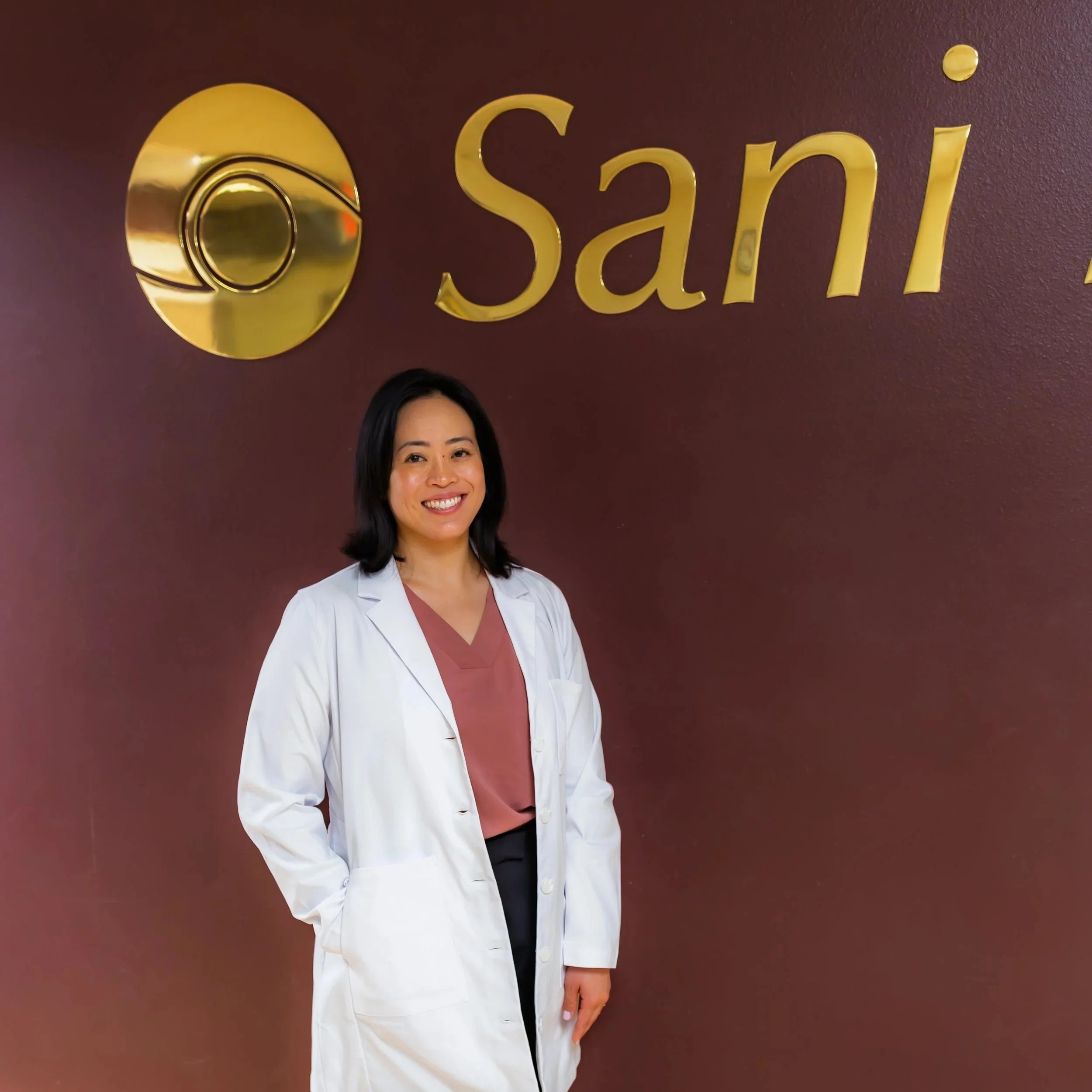 A woman with shoulder-length black hair and a white lab coat smiling and standing in front of a maroon wall with gold 3D lettering that spells 'Sani' and a gold circular logo.