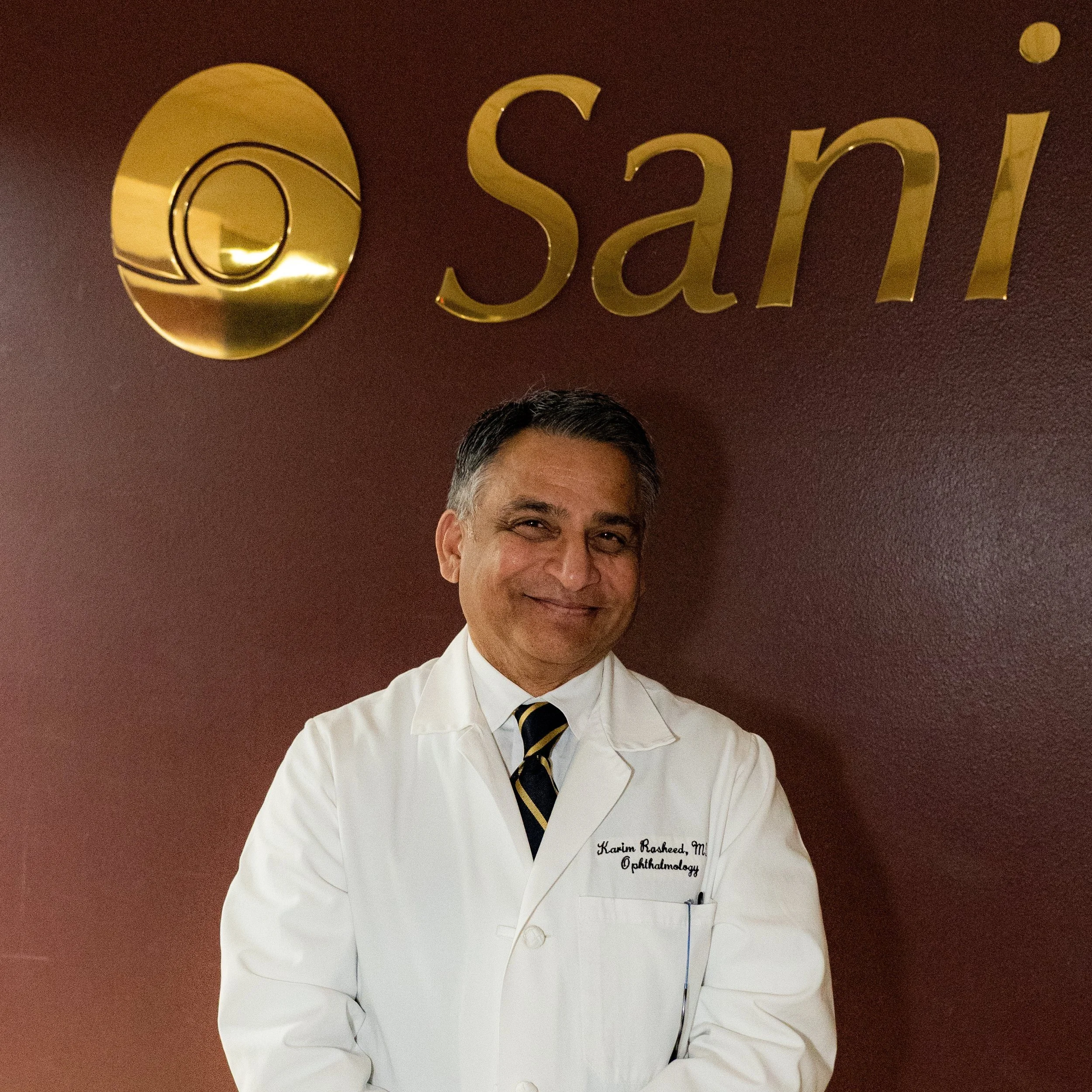 Ophthalmologist in a white coat smiling in front of the gold Sani Eye Center sign in the Templeton office.