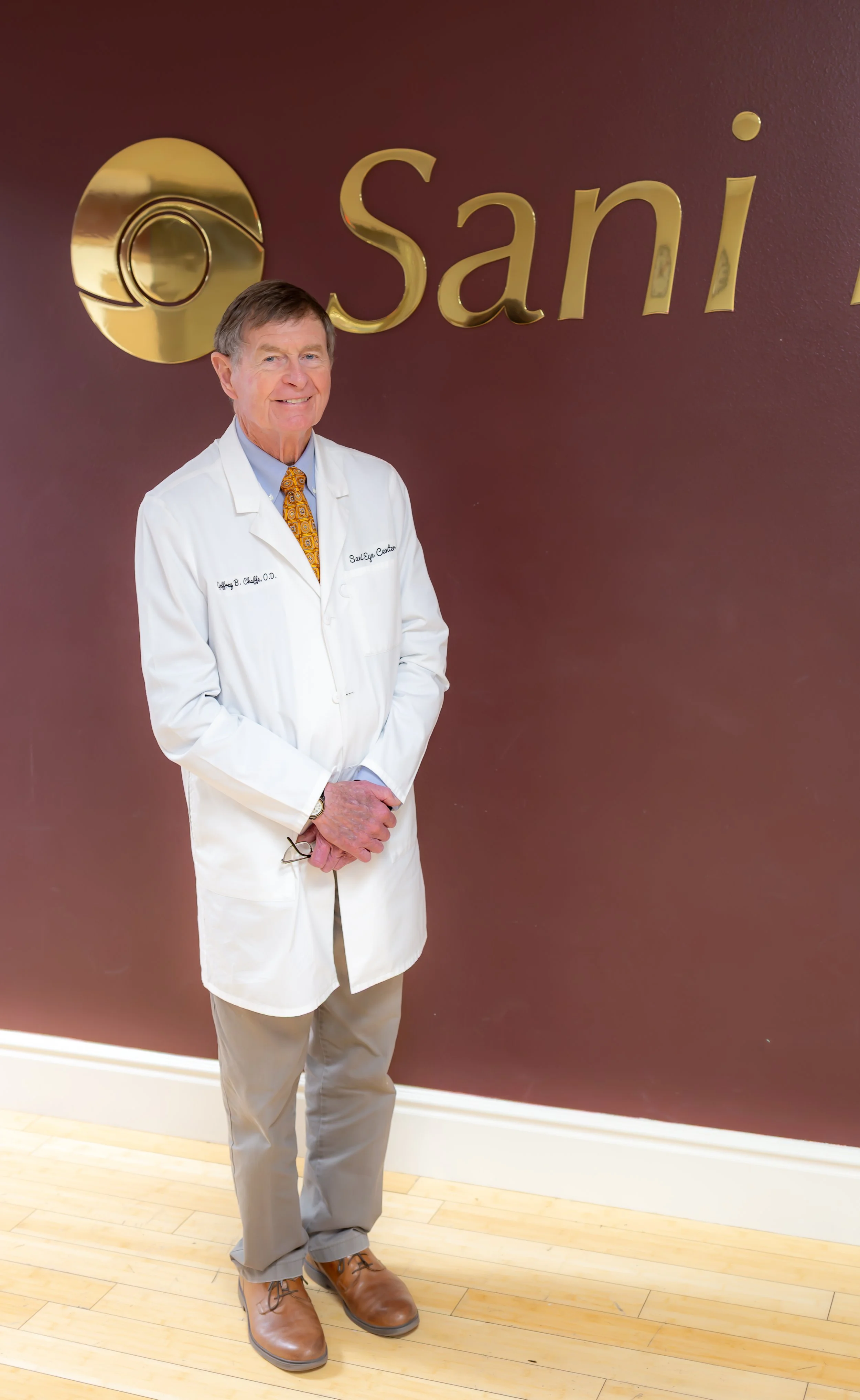 Optometrist in a white coat holding a pair of glasses, standing in front of the Sani Eye Center sign.