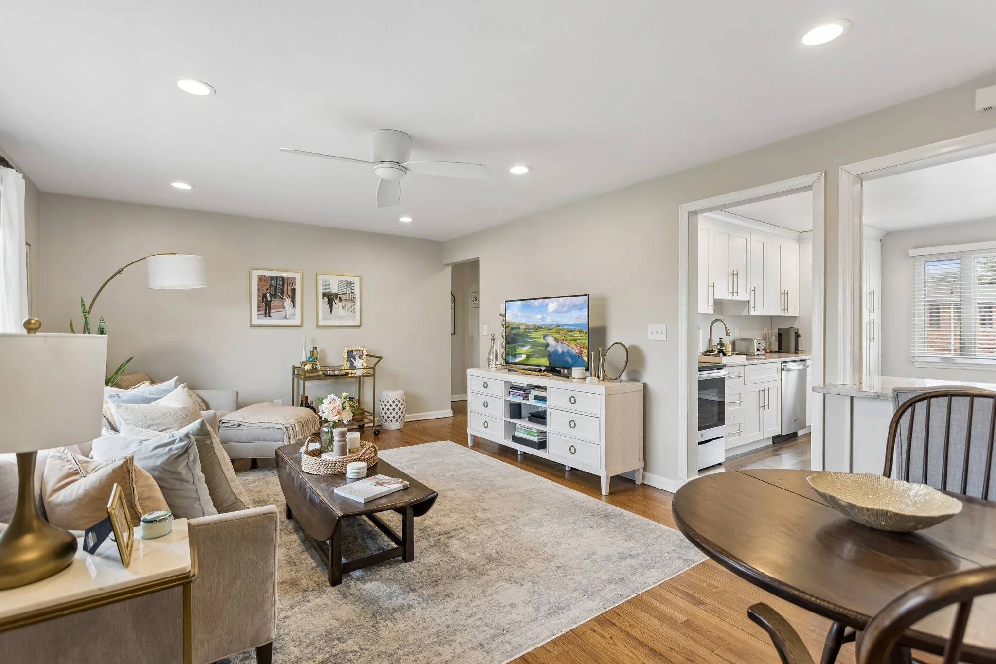 Living room with beige sofa, wooden coffee table, TV on white console, and open kitchen with white cabinets and window blinds.