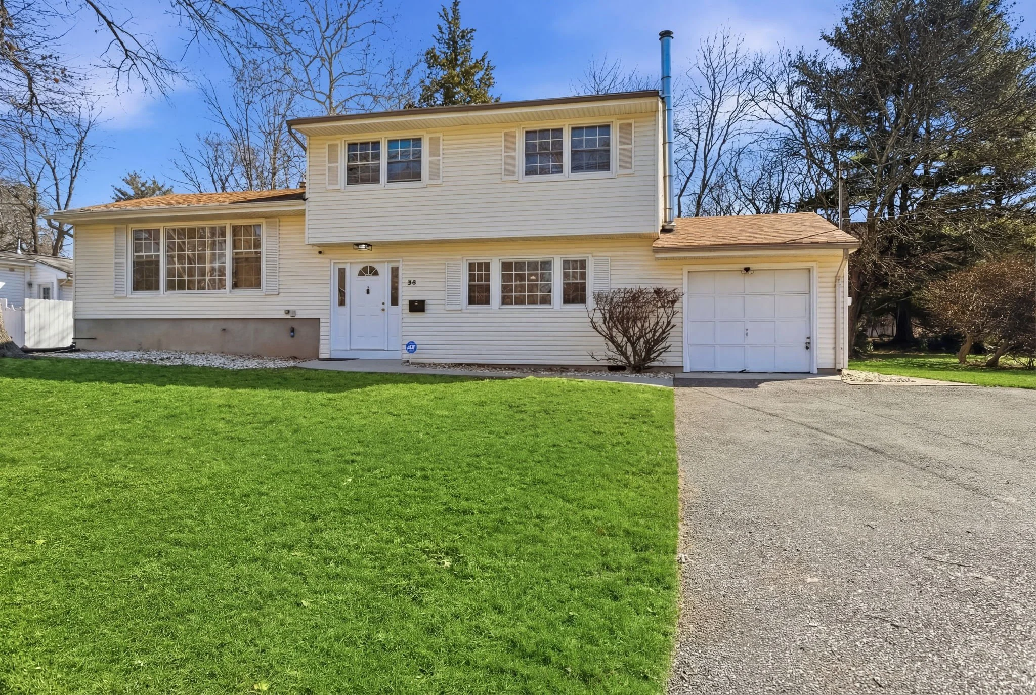 A two-story house with white siding and a brown shingled roof, with a small front lawn and driveway, set against a clear blue sky with leafless trees in the background.