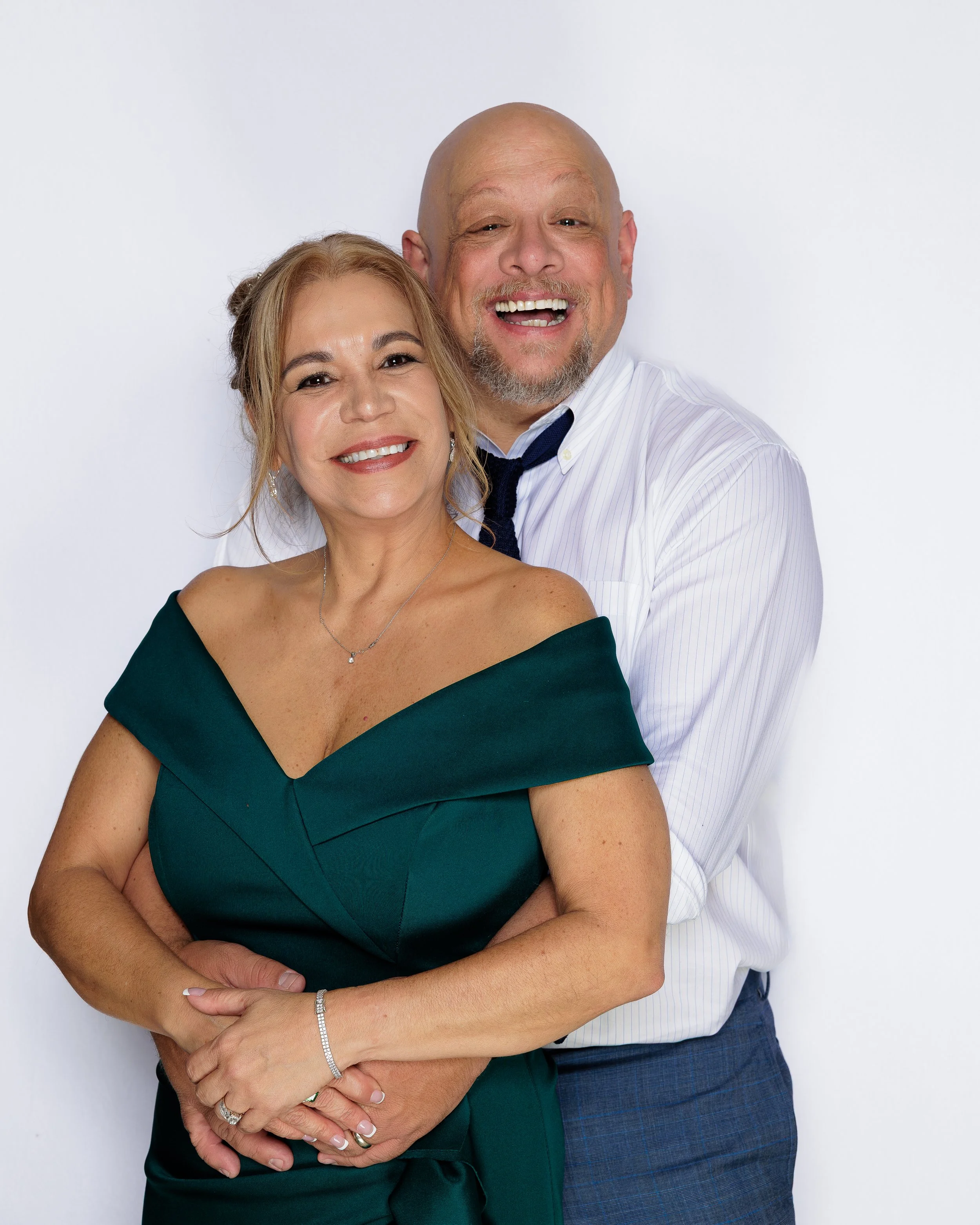 A middle-aged man and woman sharing a hug and smiling against a plain white background. The woman has light skin, shoulder-length blonde hair, and is wearing a dark green off-shoulder dress and jewelry. The man has light skin, a bald head, gray beard