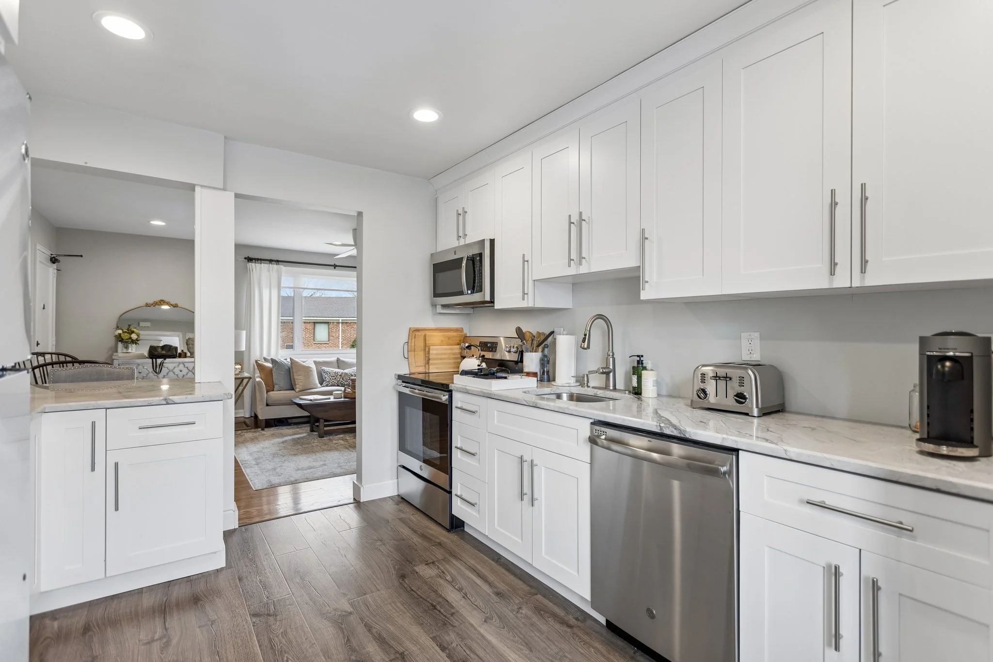 Modern white kitchen with marble countertops, stainless steel appliances, and wooden flooring, opening into a living room with a beige sofa and large windows.