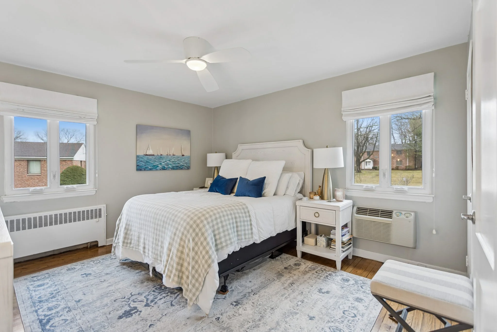 A bedroom with a white bed and blue pillows, white bedside tables with lamps, a painting of sailboats, windows with white blinds, a ceiling fan, and a patterned area rug.