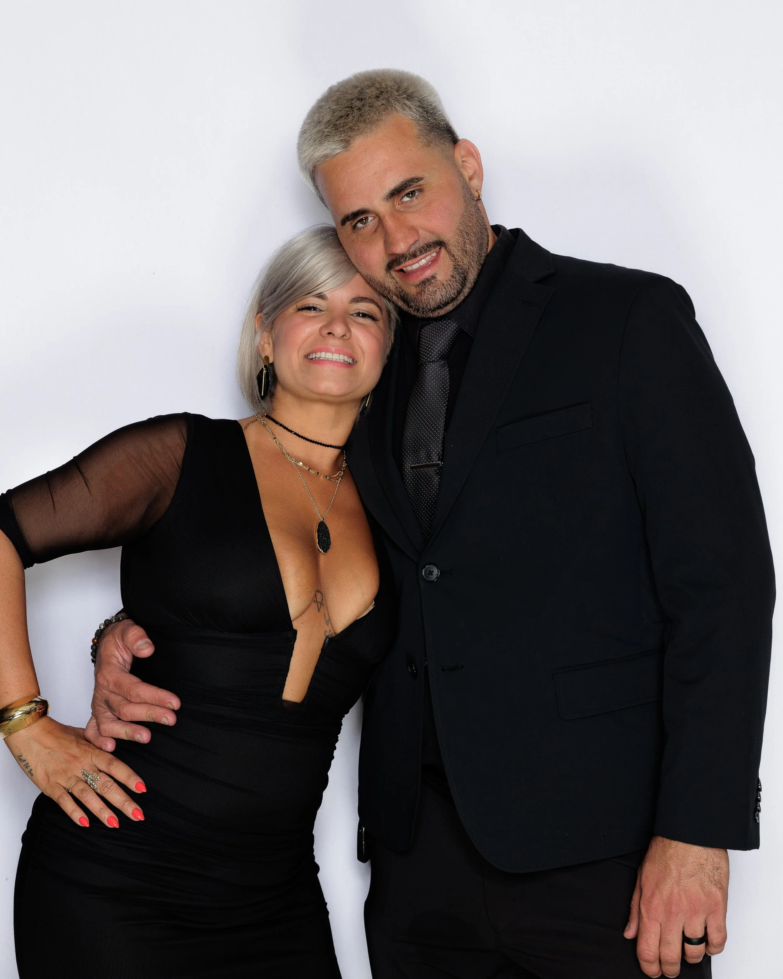 A man and woman pose closely together against a plain white background, smiling and looking at the camera. The woman has silver hair, wears a black dress with mesh sleeves, and various jewelry. The man has short gray hair, wears a black suit, and has