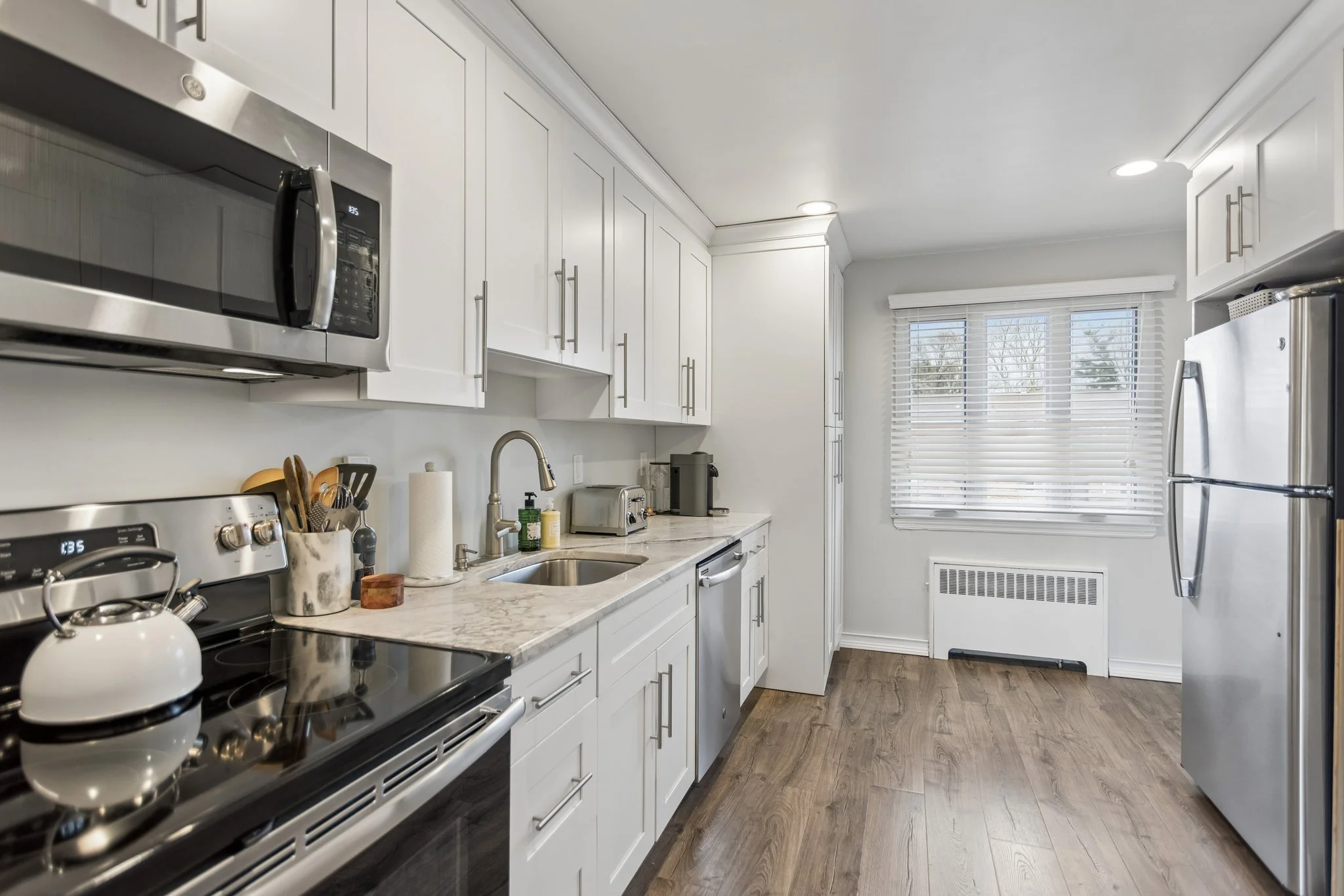 A modern kitchen with white cabinets, stainless steel appliances, a marble countertop, a window with blinds, and a wood floor.