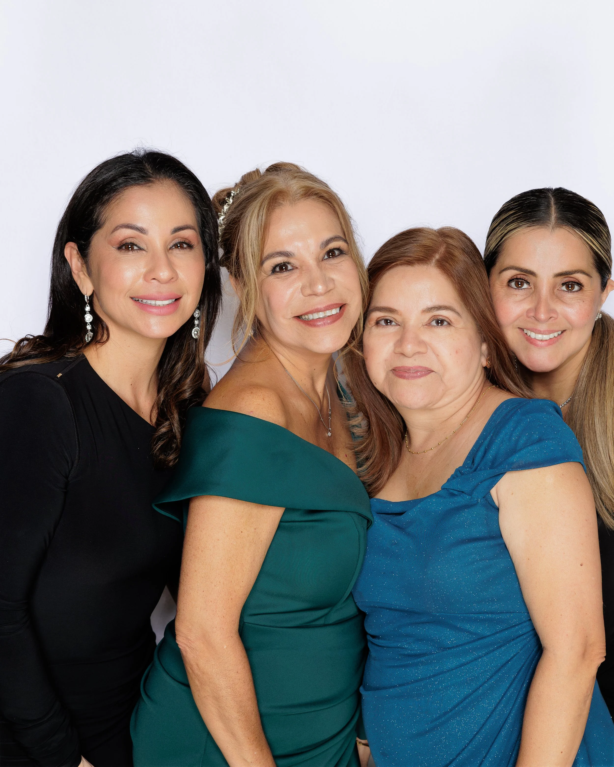Four women closely huddled together and smiling at the camera, each wearing elegant dresses and jewelry, against a plain white background.