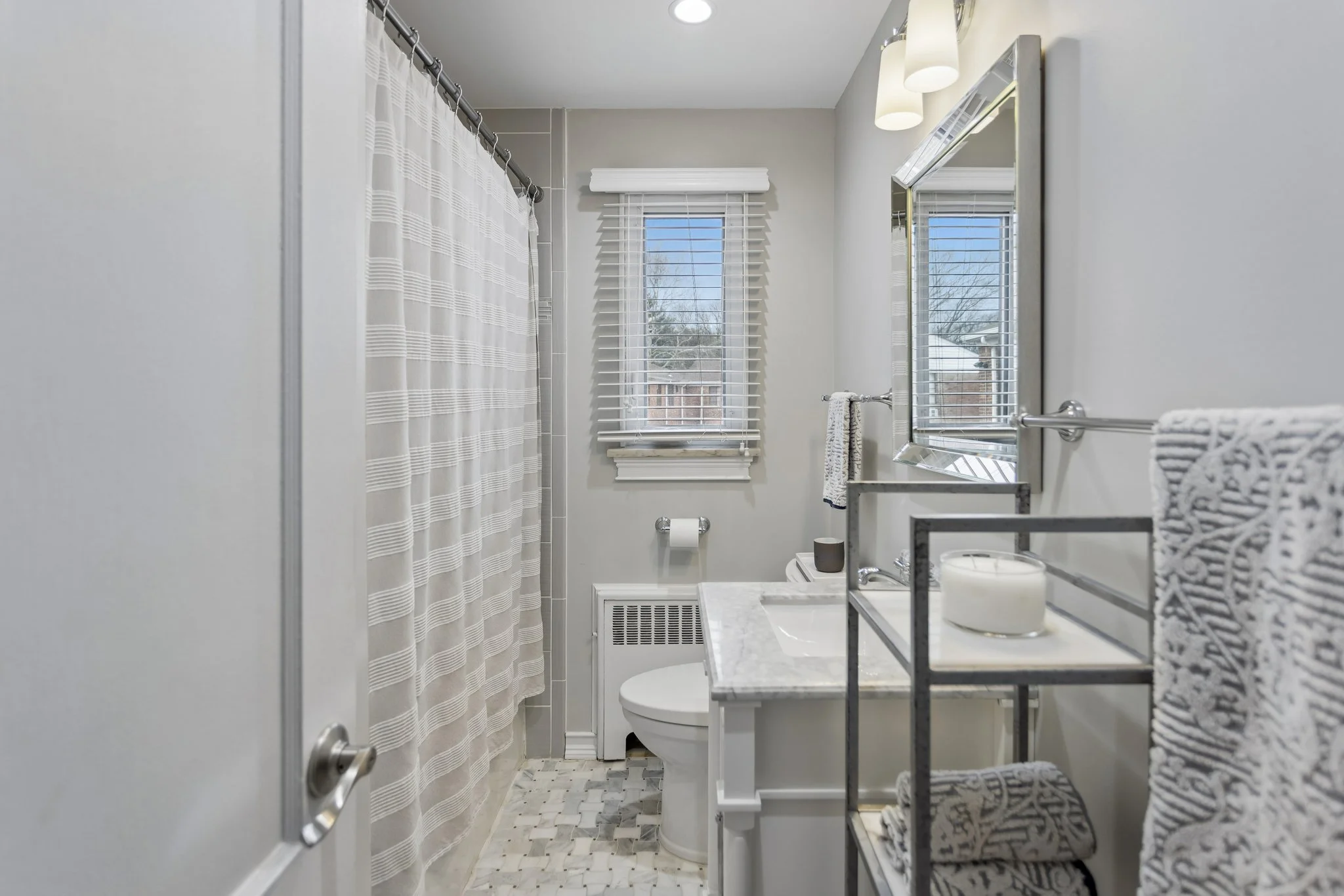 A small bathroom with a window above the toilet, a light-colored shower curtain, and various toiletries on a metal rack, with a mirror and light fixtures above the sink.
