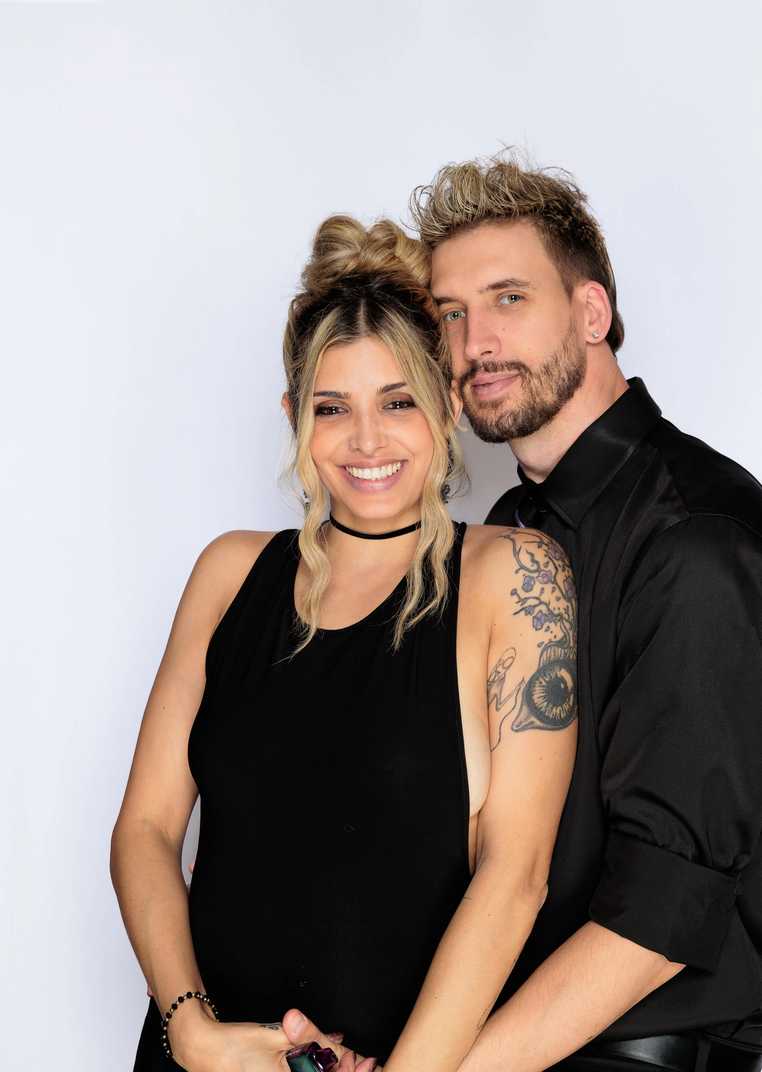 A smiling woman in a black sleeveless top with tattoos on her left arm, and a man in a black shirt with styled hair, posing close together against a plain white background.