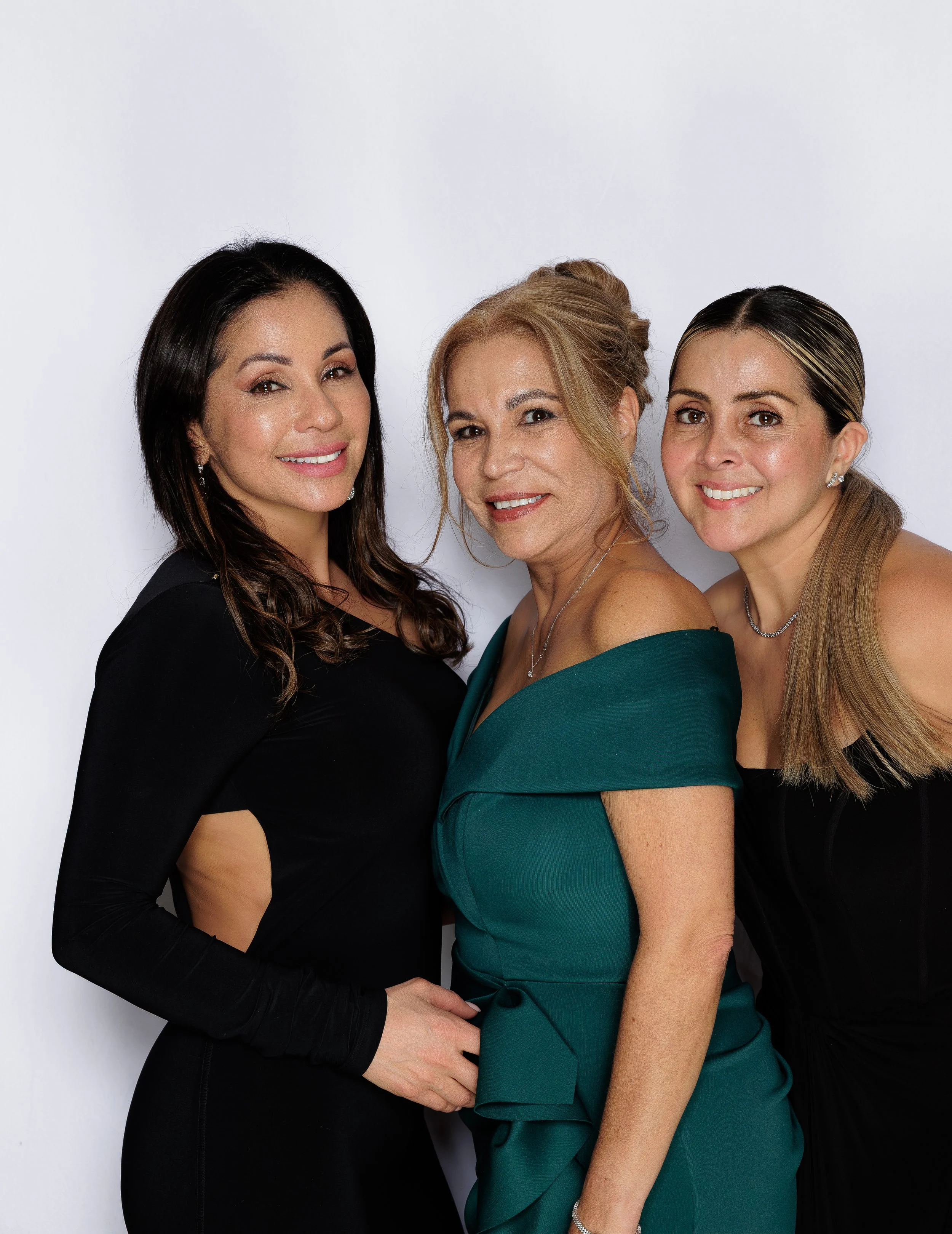 Three women standing close together against a plain white background, smiling at the camera. The woman on the left has long dark hair and is wearing a black dress with long sleeves. The woman in the middle has blonde hair styled up, wearing an off-sh