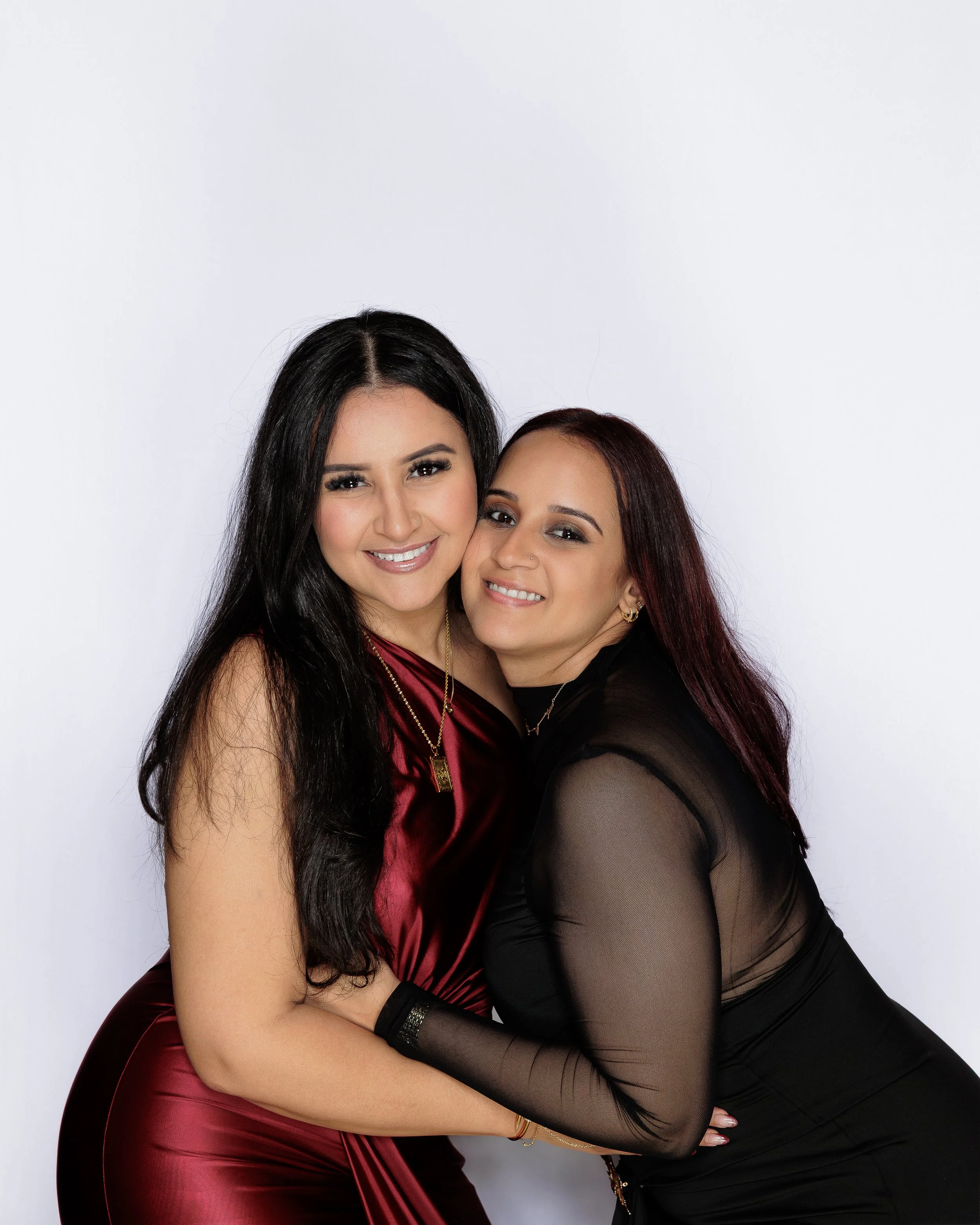 Two women with dark brown hair and light to medium skin tones embrace and smile at the camera. The woman on the left wears a sleeveless, burgundy satin dress, while the woman on the right wears a black sheer top with long sleeves and matching black p