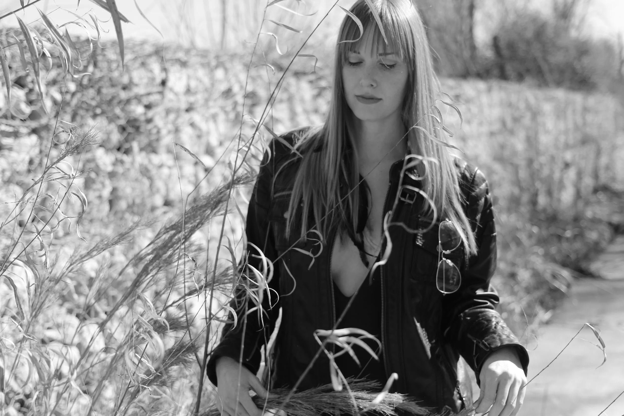A woman with long hair wearing a leather jacket, walking outdoors among tall grass