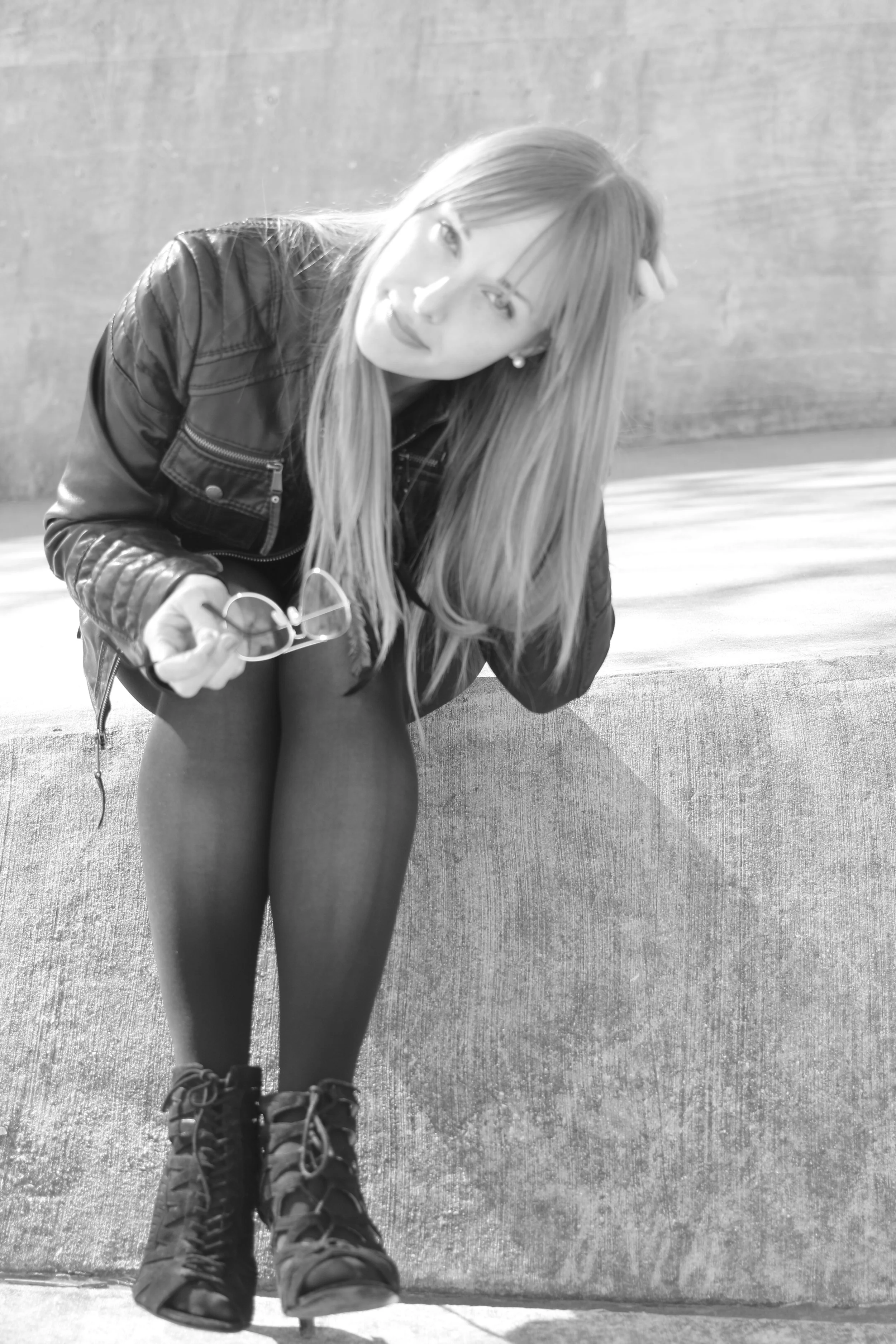 A young woman with long hair sitting on a concrete ledge, holding glasses in her hand and looking at the camera, wearing a leather jacket, tights, and combat boots.