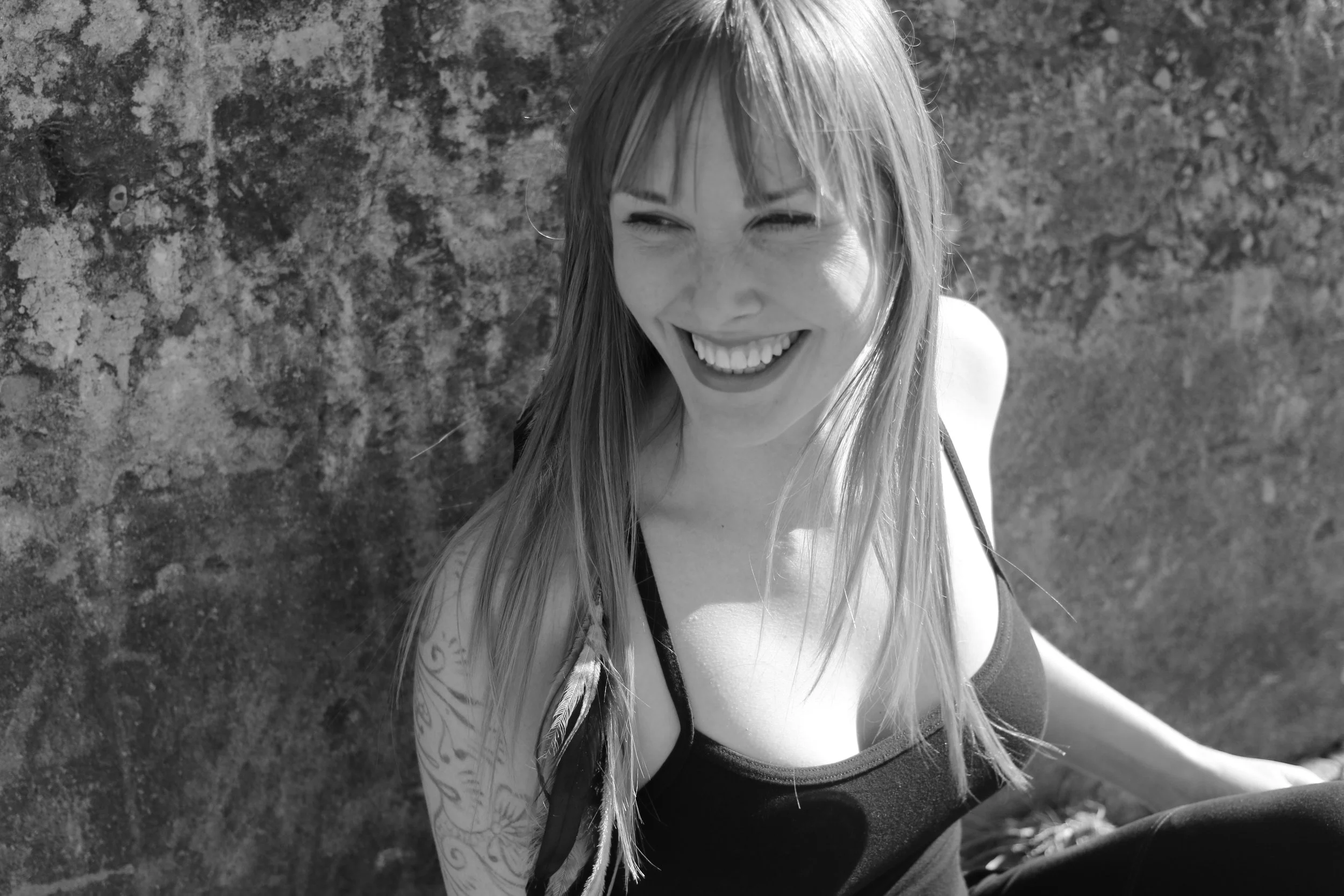 A woman with long hair and a tattoo on her arm is smiling and sitting outdoors near a textured stone wall.