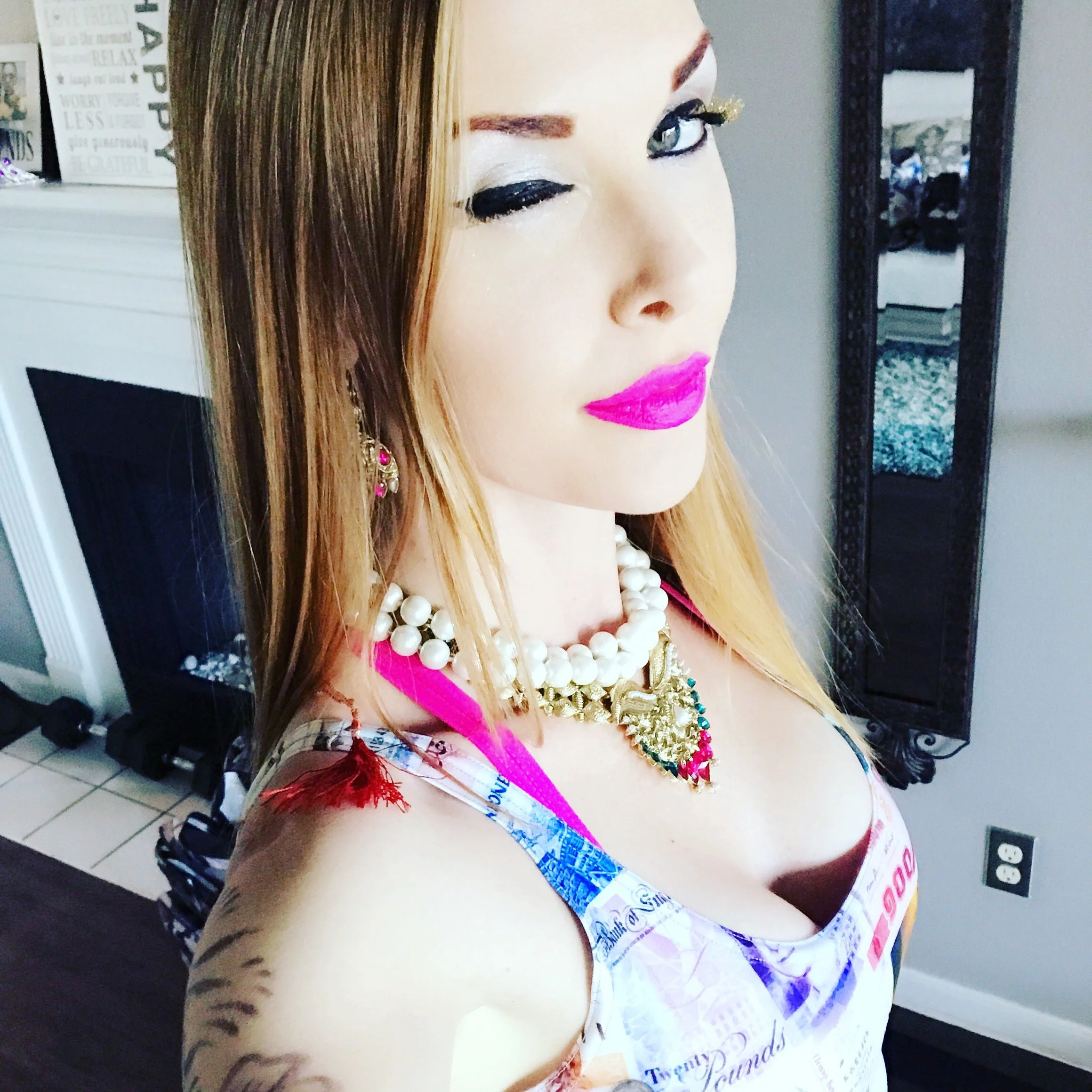 A woman with long blonde hair, wearing vibrant pink lipstick and eye makeup, dressed in a top made of newspaper print, accessorized with pearl and gold necklaces and earrings, standing in an indoor setting.