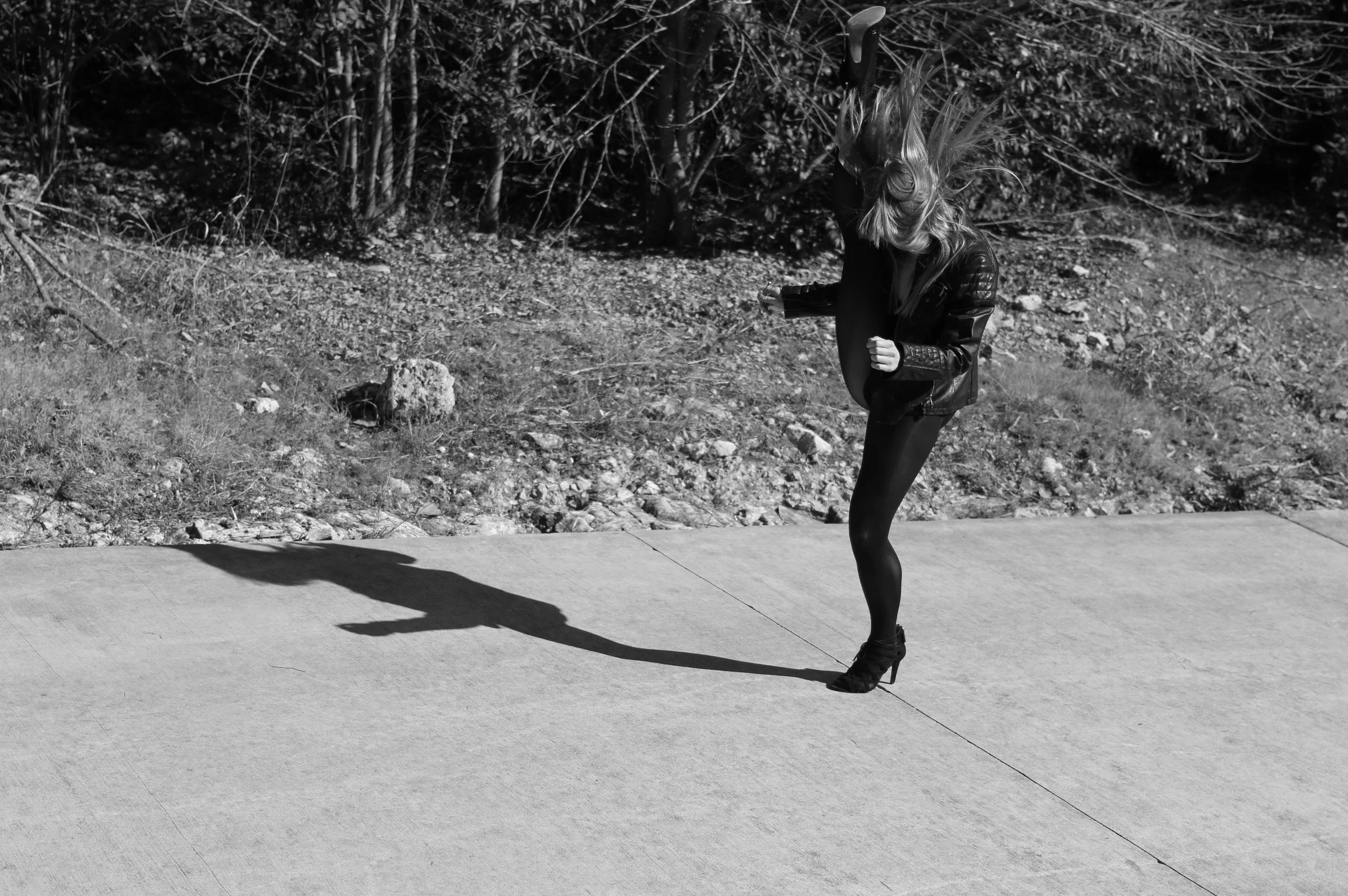 Black and white photo of a woman with hair flying, standing on a sidewalk with a large shadow cast on the ground, holding a skateboard, with trees and rocks in the background.