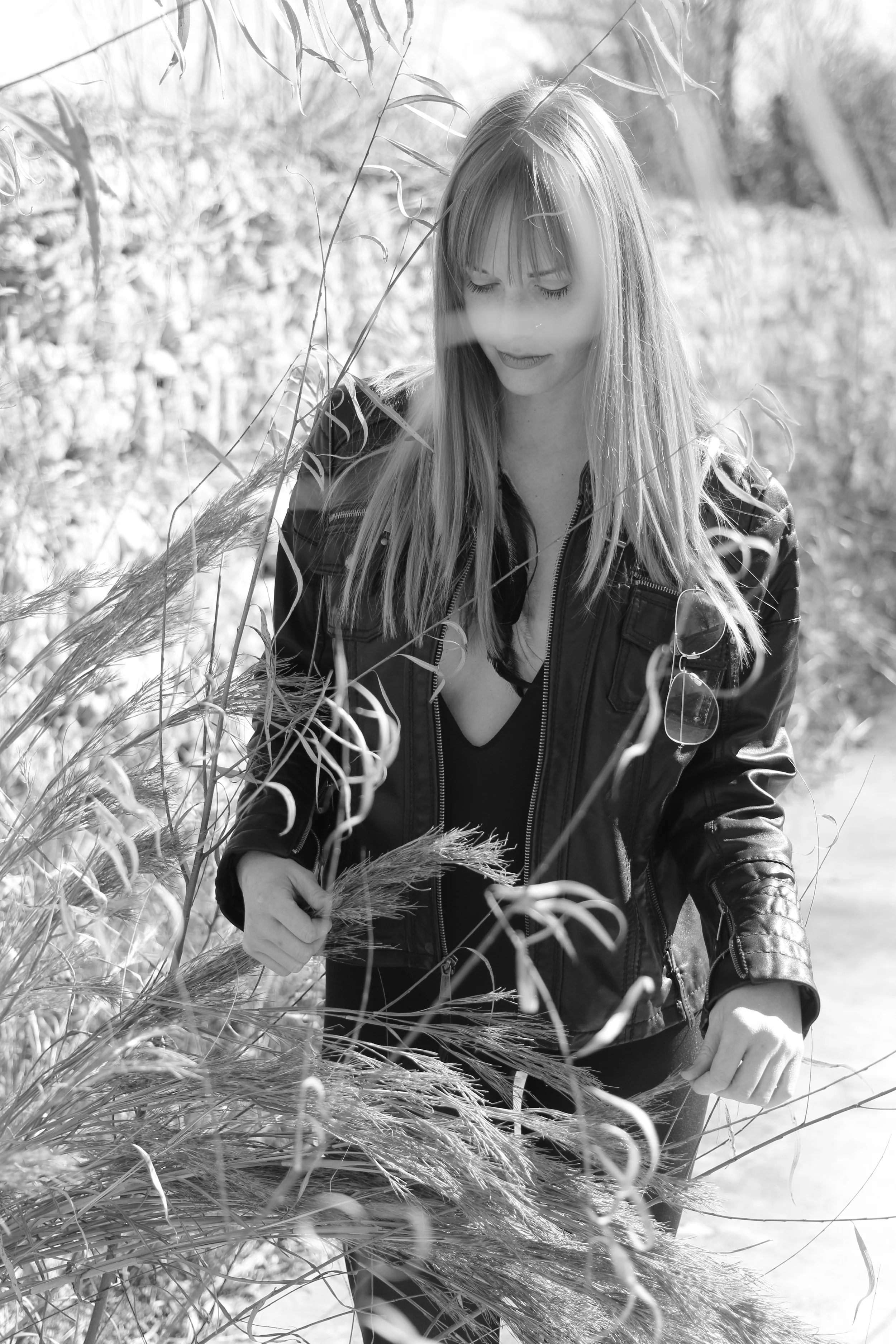 A woman with long hair wearing a leather jacket and sunglasses hanging from it, walking through tall grass or weeds outdoors on a bright day.