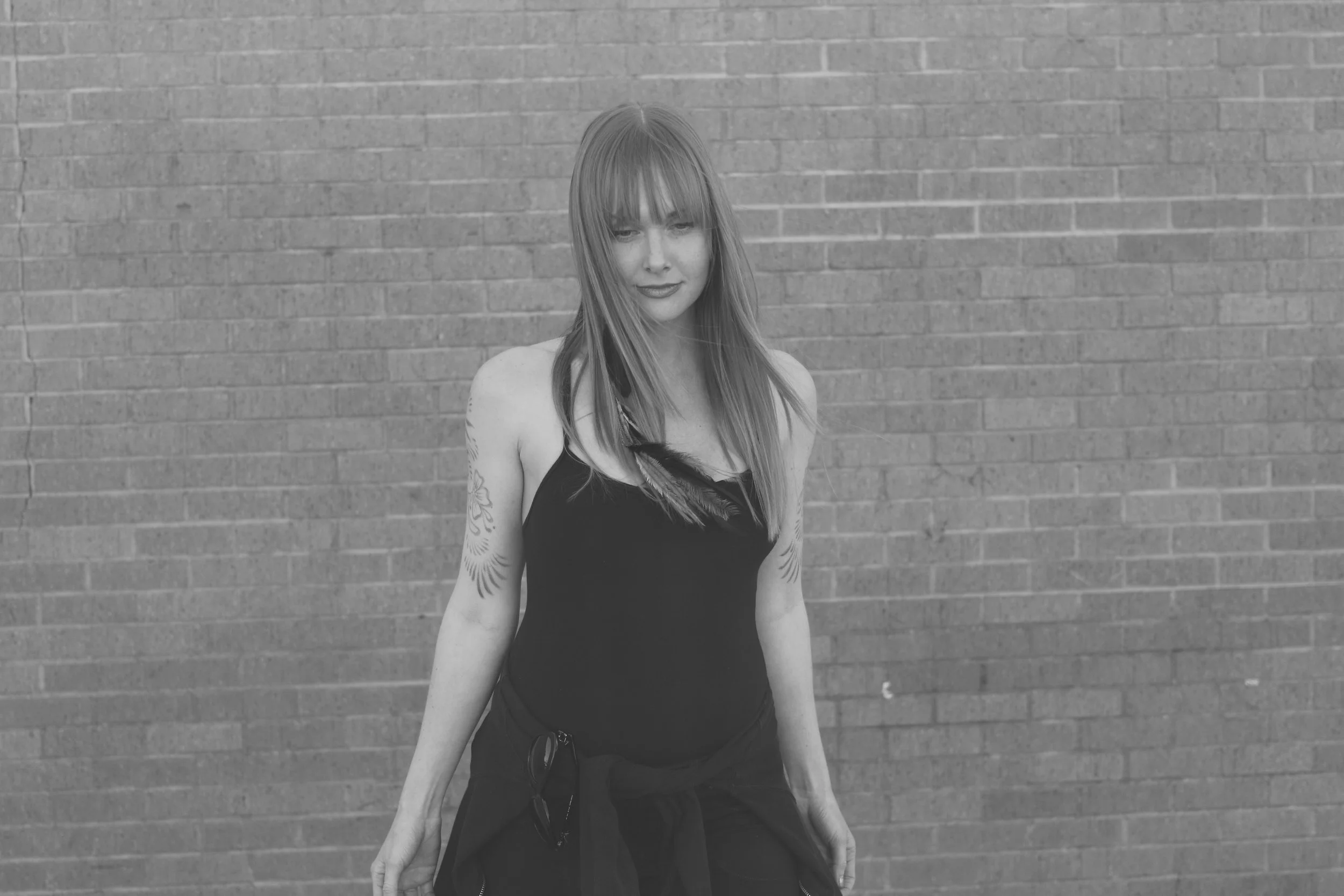 A woman with straight hair, wearing a black tank top, standing against a brick wall. She has tattoos on both arms and glasses hanging from her waistband.