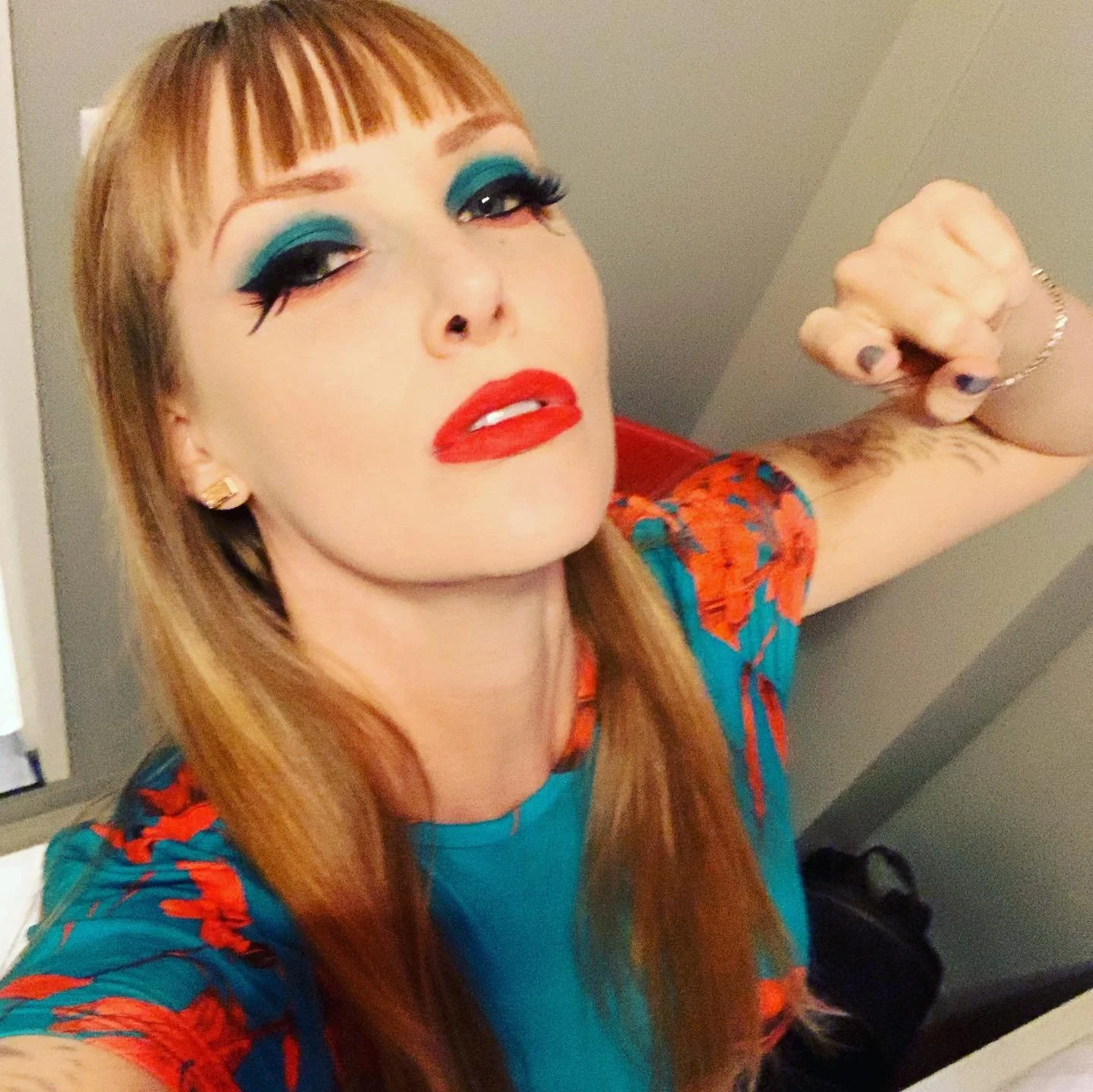 A woman with long red hair, bold makeup with blue eyeshadow, black eyeliner, and red lipstick, looking directly at the camera with a confident expression. She is wearing a colorful top with visible floral patterns and has a tattoo on her forearm. She