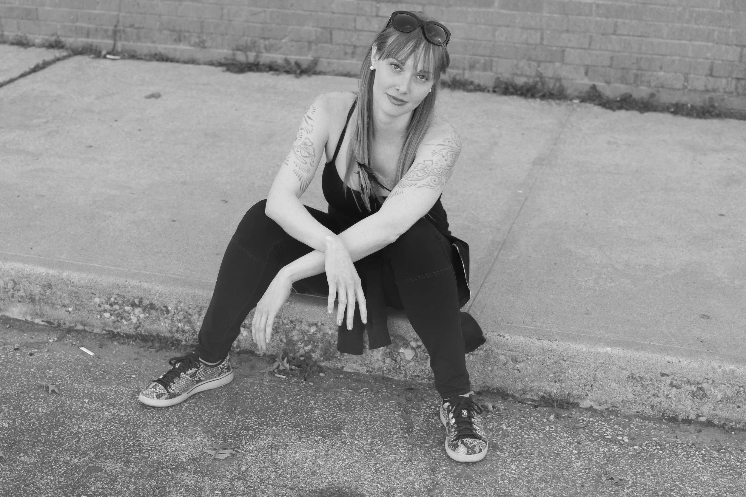 A woman with tattoos on her arms sitting on a curb outside, wearing sunglasses on her head and sneakers.