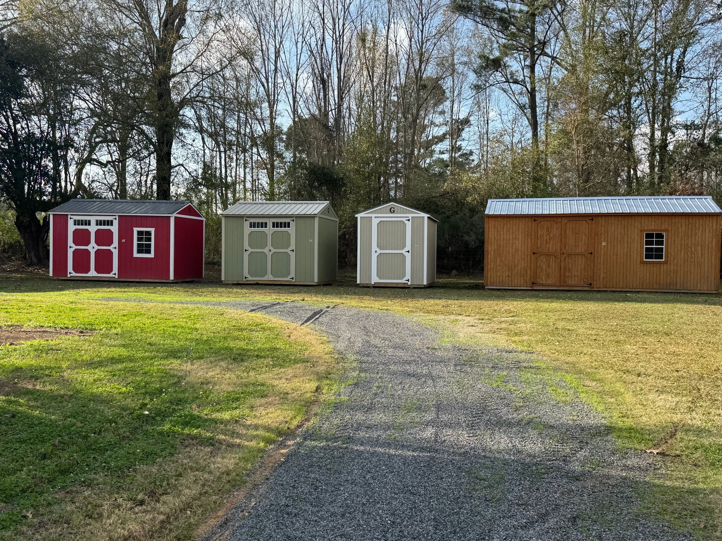 Four garden sheds on a grassy lawn with a gravel pathway. The sheds are red, green, white, and brown, with the red and white sheds having double doors, and the brown shed having sliding doors and a small window.