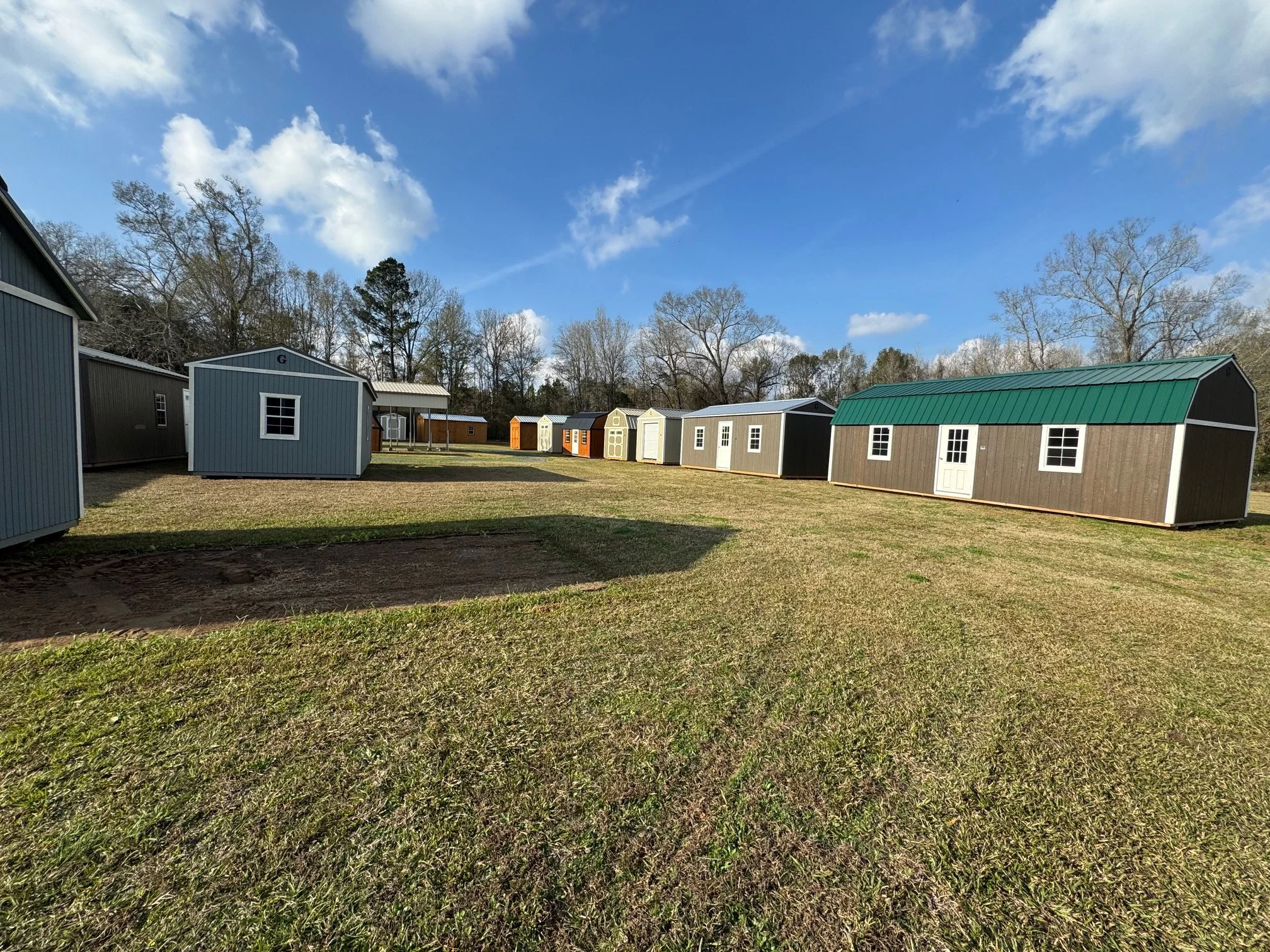Multiple small storage sheds of various colors and styles in a grassy area under a partly cloudy sky.