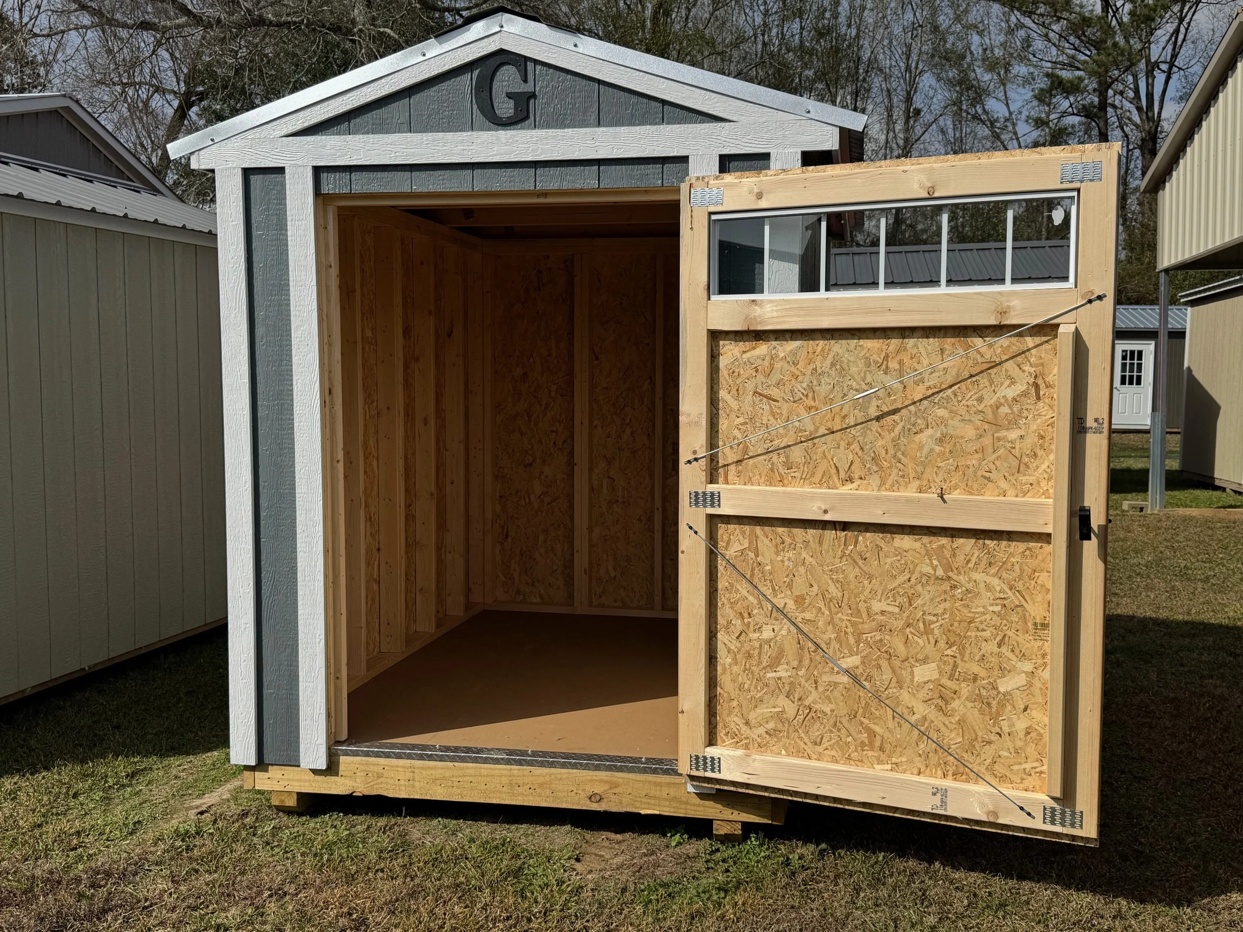 A small wooden shed under construction with an open door, showing the interior framing and windows on the upper front wall.