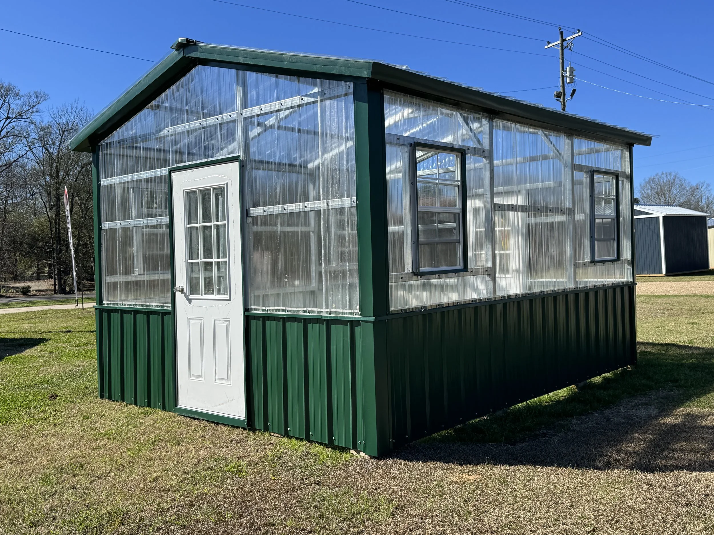 Greenhouse with transparent walls and a white door, situated on a grassy lawn under a clear blue sky.