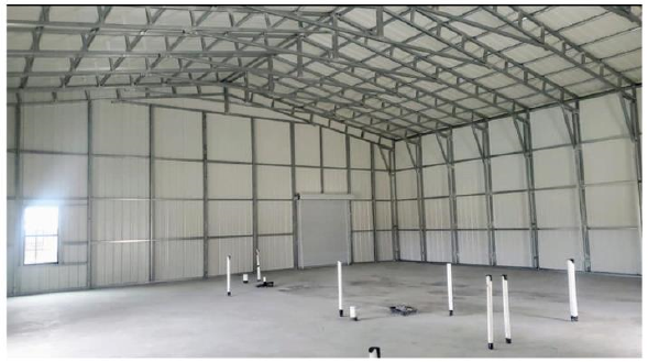 Empty industrial warehouse or hangar with metal walls, ceiling, and floor, with white poles arranged on the ground and a closed garage door on the back wall.