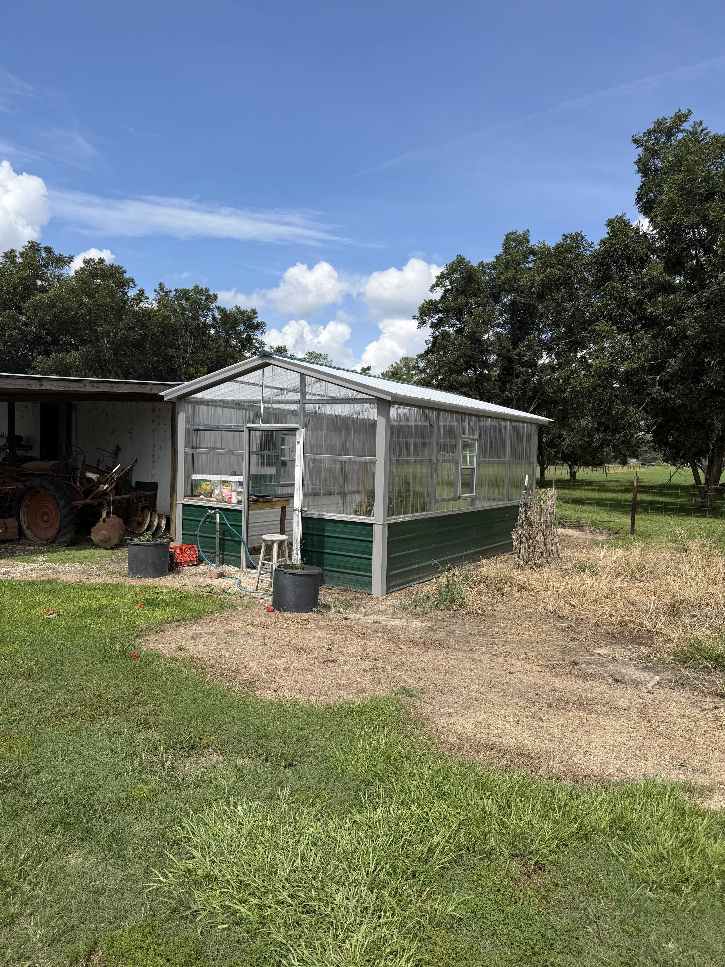 A greenhouse with a metal roof and transparent walls on a farm, with green trees behind and a partly cloudy sky above.