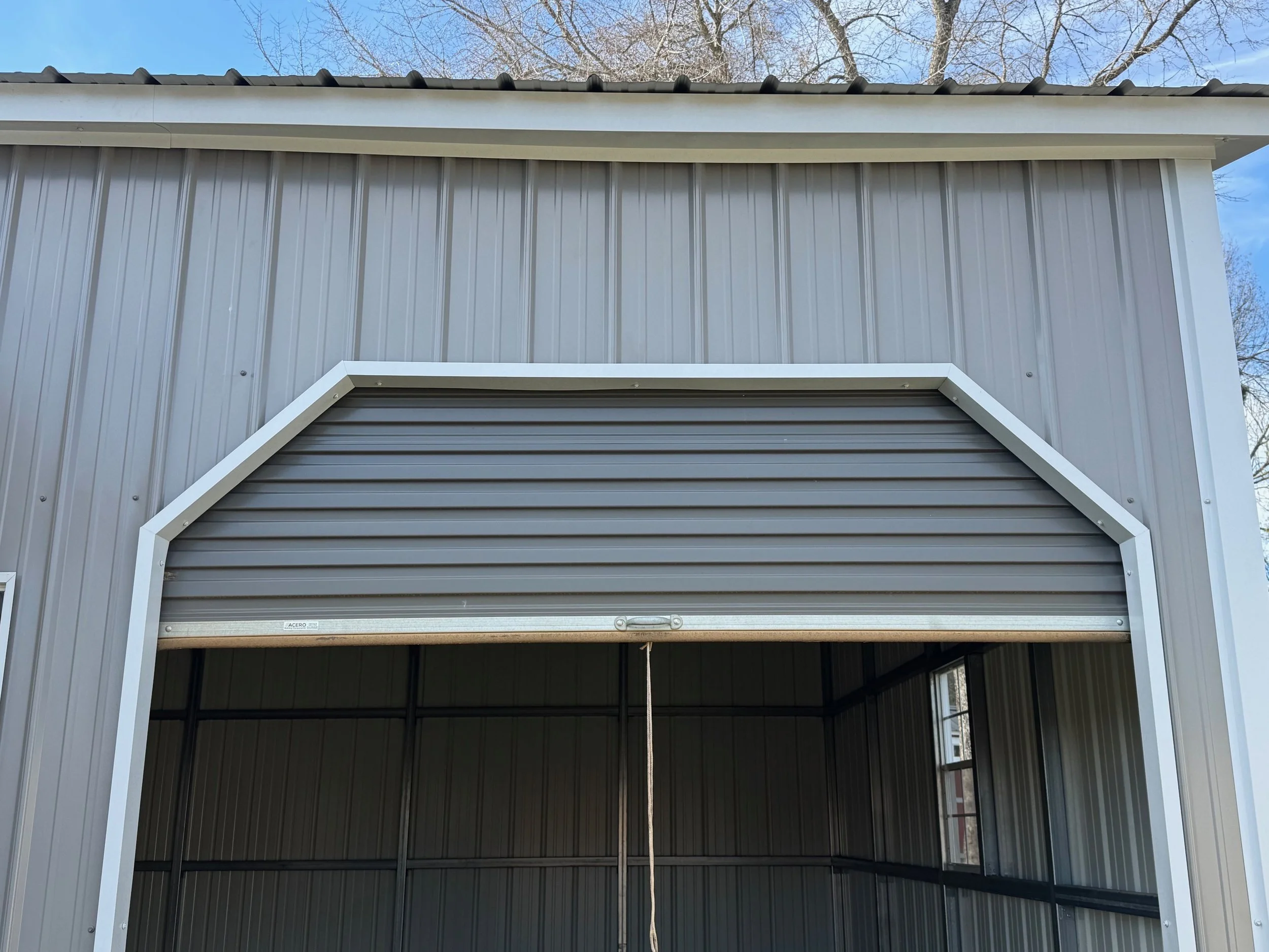 Front view of a metal storage shed with a partially open roll-up door, gray siding, and a sloped roof, with trees and a clear blue sky in the background.