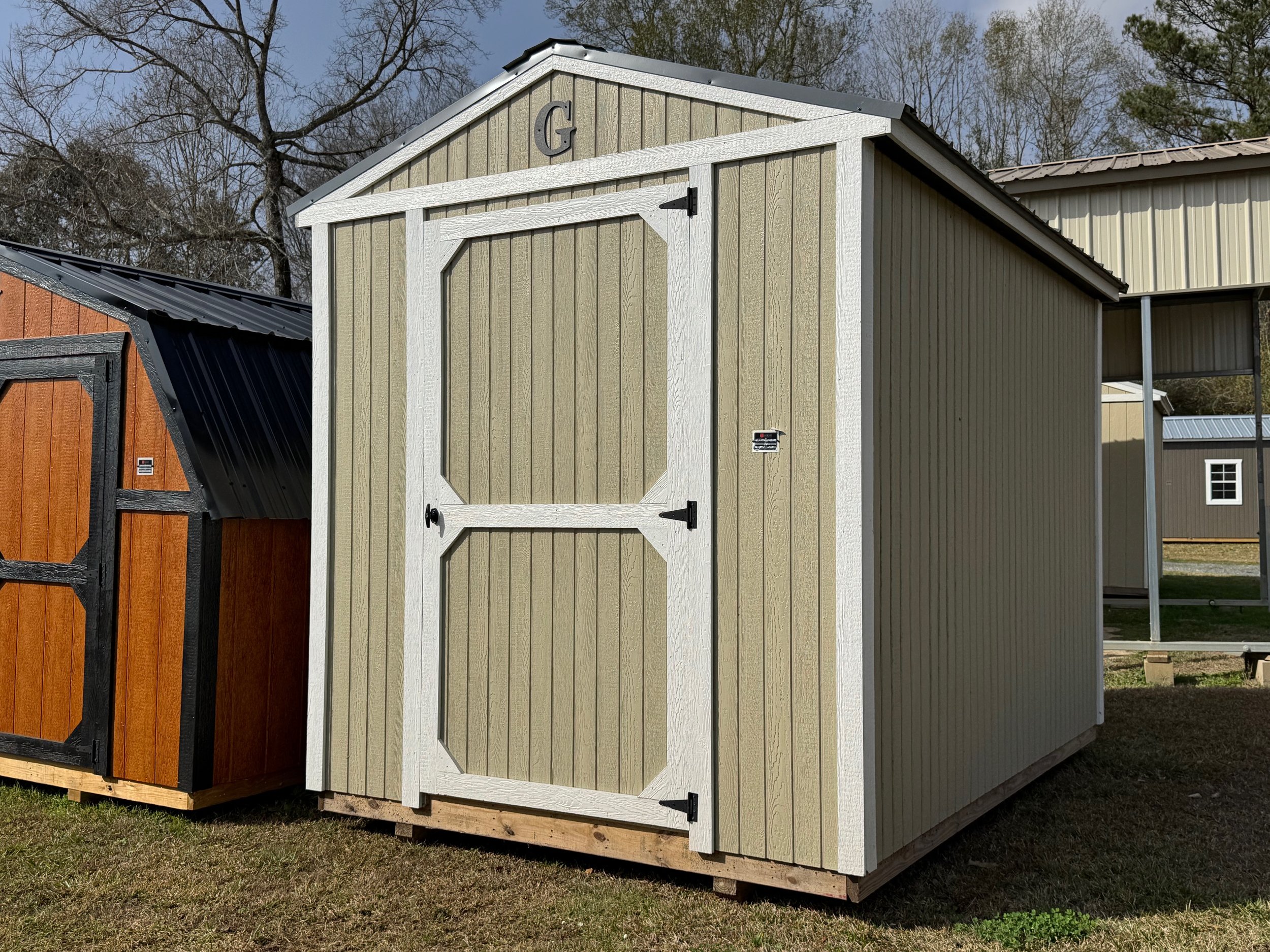 A small beige outdoor shed with white trim and a gable roof, situated on a grassy area, with neighboring sheds visible in the background.