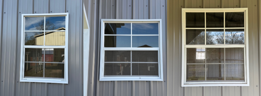 Three double-hung windows on a metal building wall, reflecting the sky and trees.